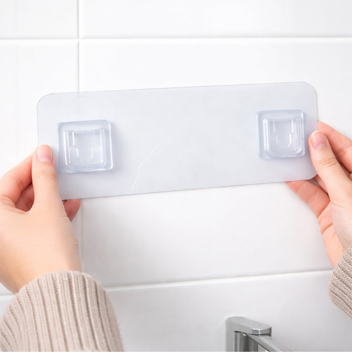 Hands position a clear plastic self-adhesive hanger with two hooks on a white tiled bathroom wall.