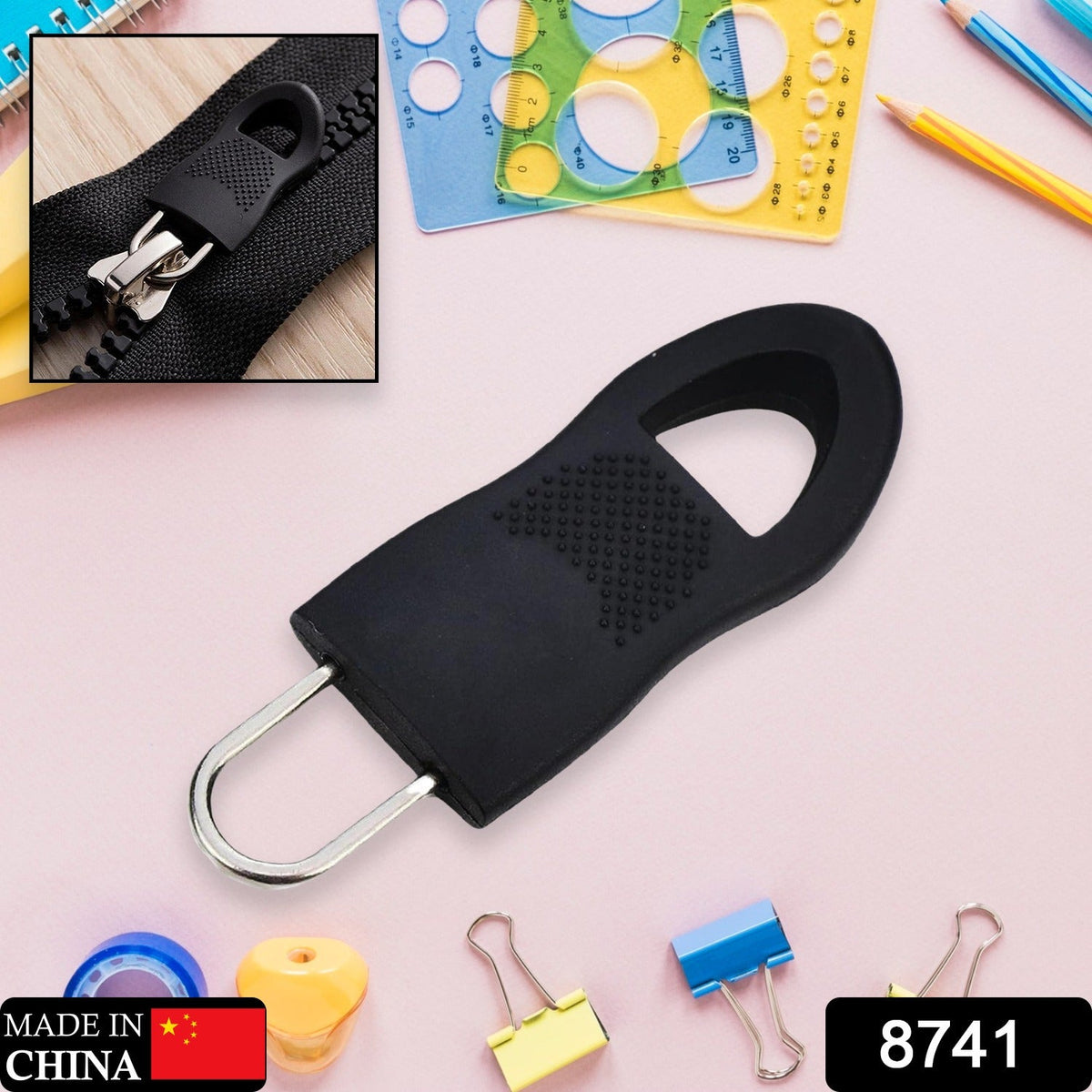 A black rubber zipper pull tab with a silver metal loop is centered on a pink surface with school supplies.