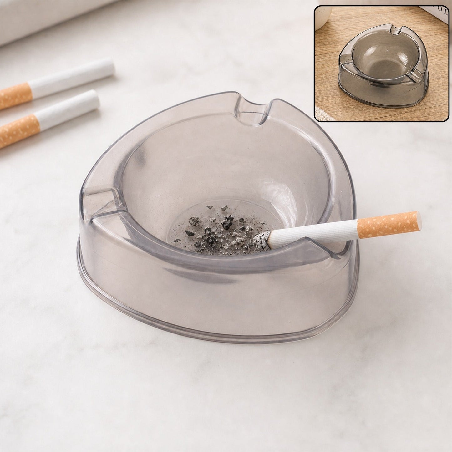 A transparent plastic ashtray with a lit cigarette and ash, plus two unlit cigarettes, rests on a light surface.