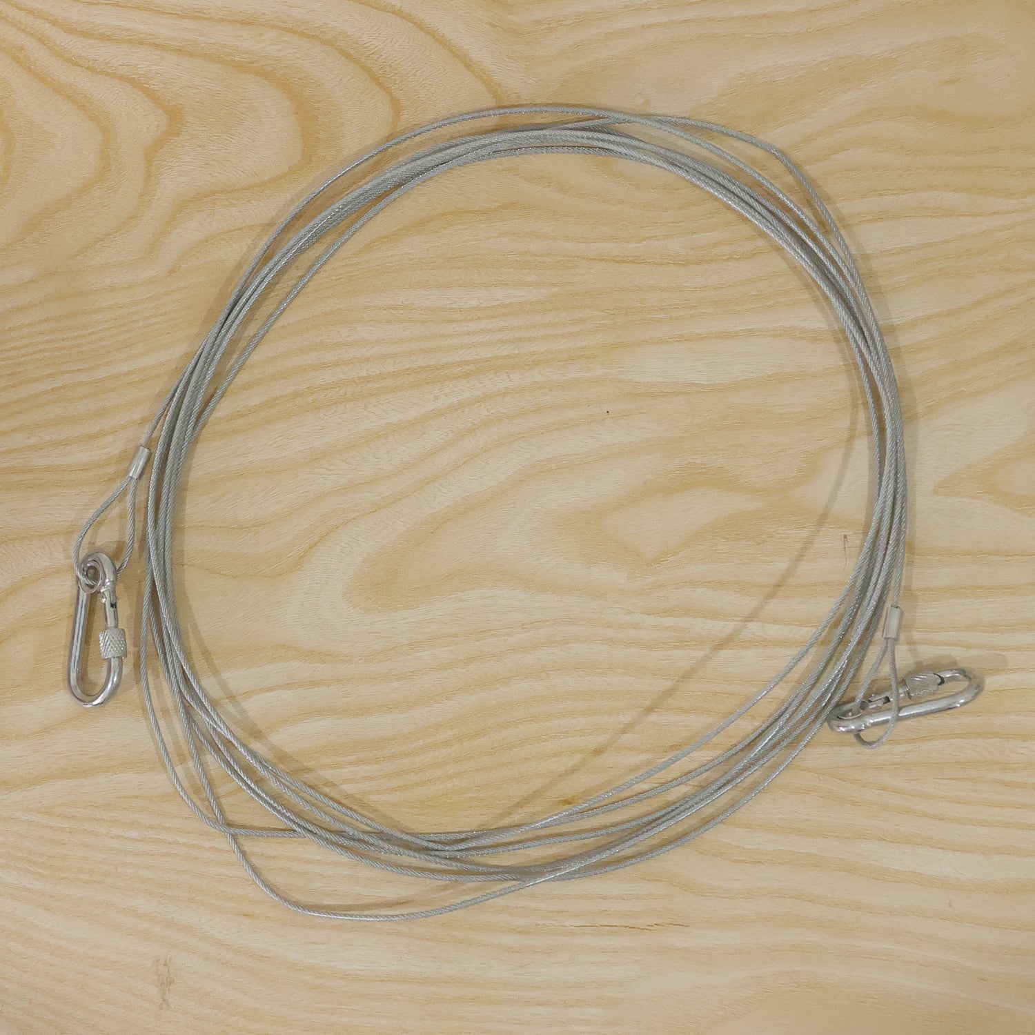 A long, silver stainless steel wire rope with two carabiner hooks is laid out on a light wooden surface.