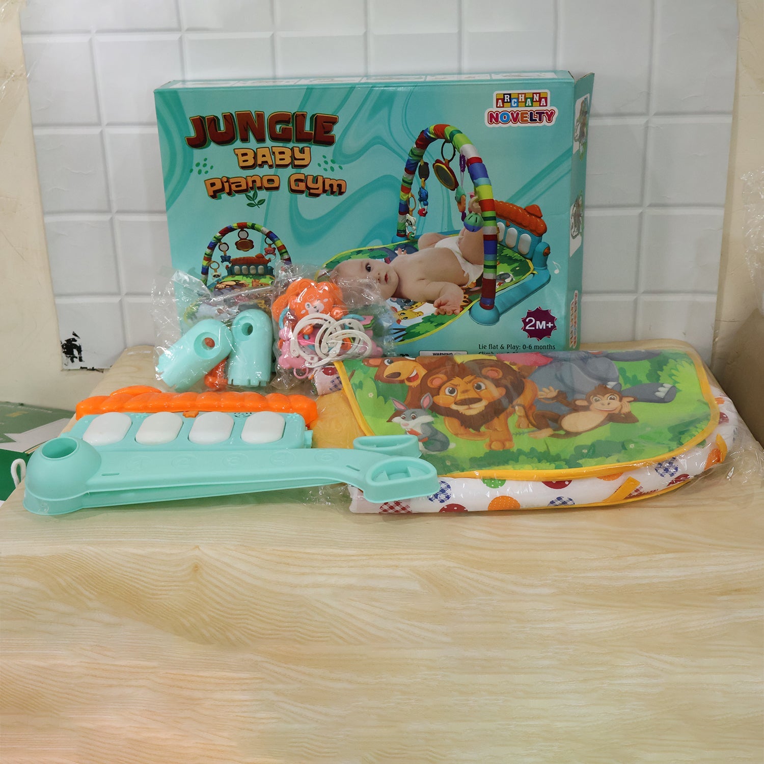 The Baby Piano Gym Activity Play Mat is shown disassembled in its packaging, with the box, mat, and piano pieces visible.