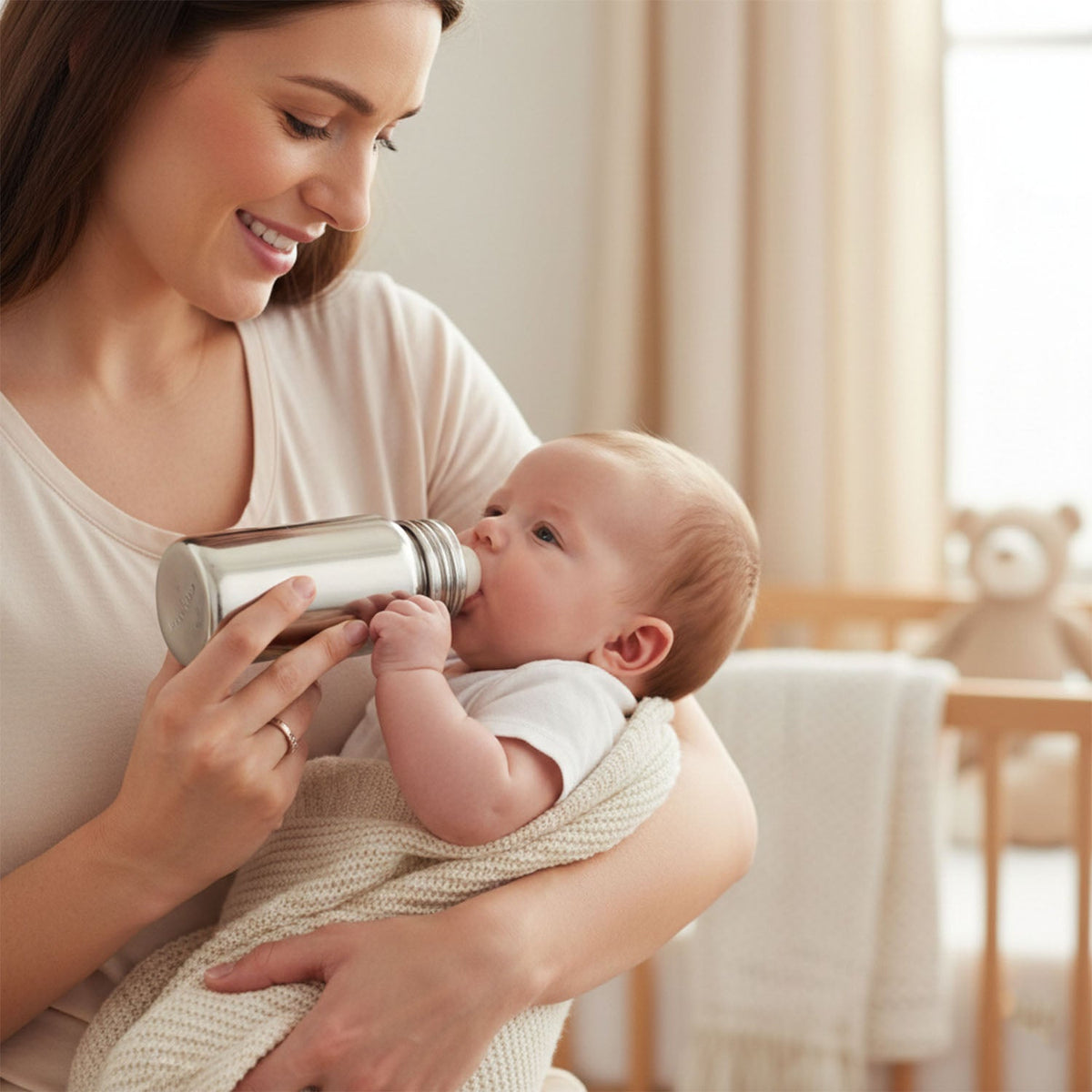 A silver stainless steel baby bottle with a clear nipple is shown next to its matching metal cap on a white background.
