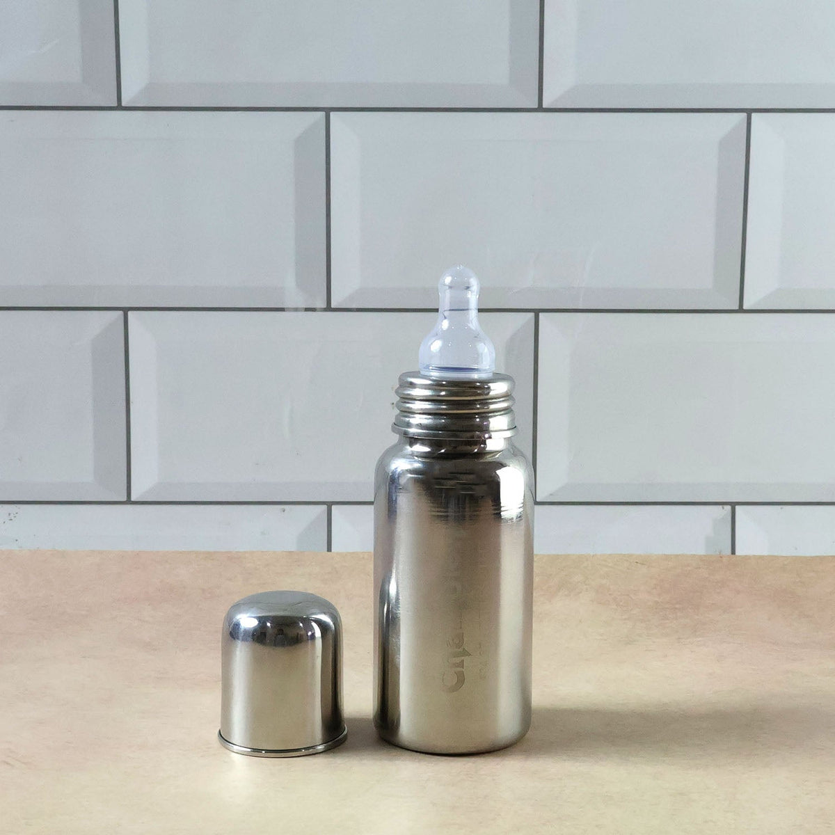 A stainless steel feeding bottle with its cap on stands beside its product box, which features an image of the bottle and