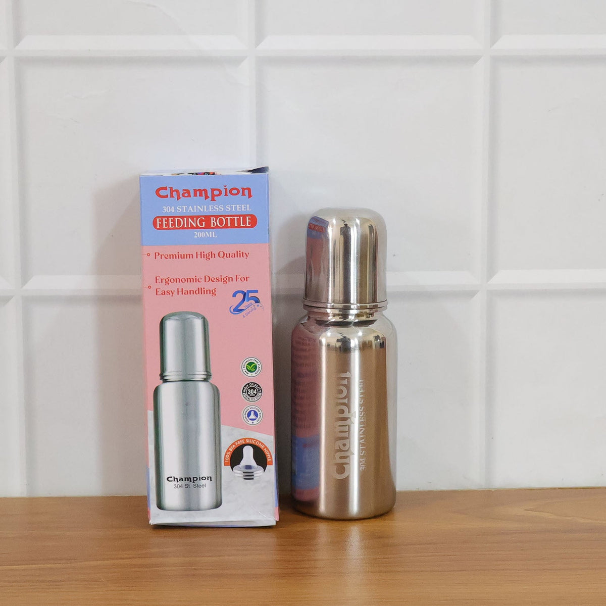 A stainless steel feeding bottle with a white nipple and its cap are shown on a wooden table, with a blurred room in the