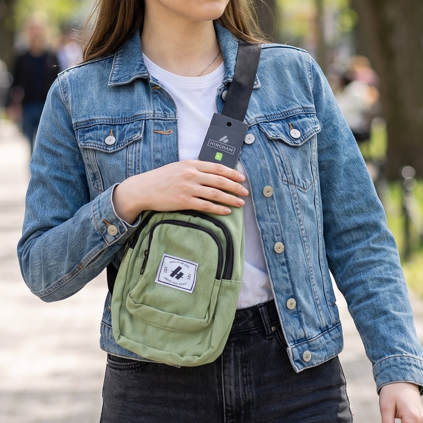 A woman in a denim jacket wears a green canvas sling bag across her chest, walking on a paved path.
