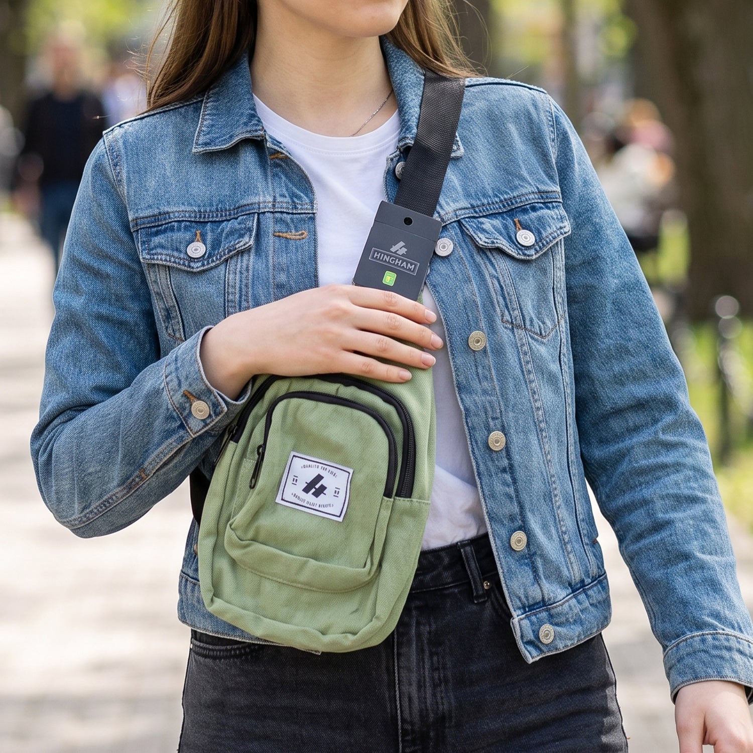 A woman in a denim jacket wears a green canvas sling bag across her chest, walking on a paved path.