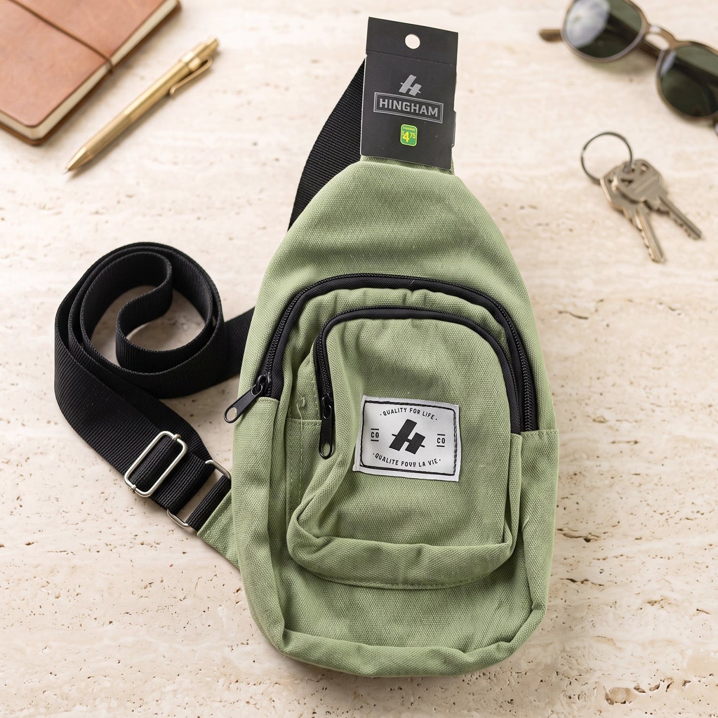 A green canvas sling bag with a black strap and front pocket lies on a textured surface next to a pen and keys.