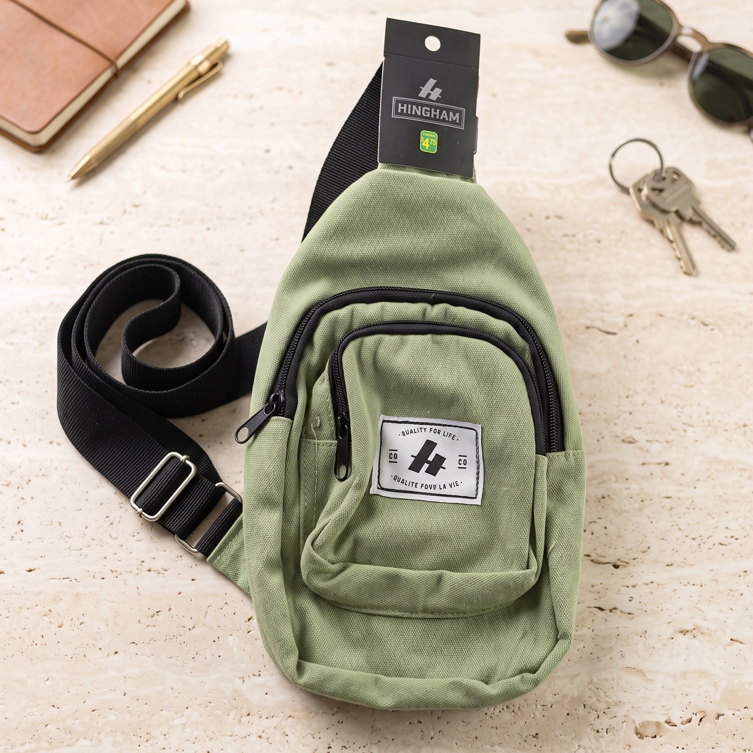 A green canvas sling bag with a black strap and front pocket lies on a textured surface next to a pen and keys.