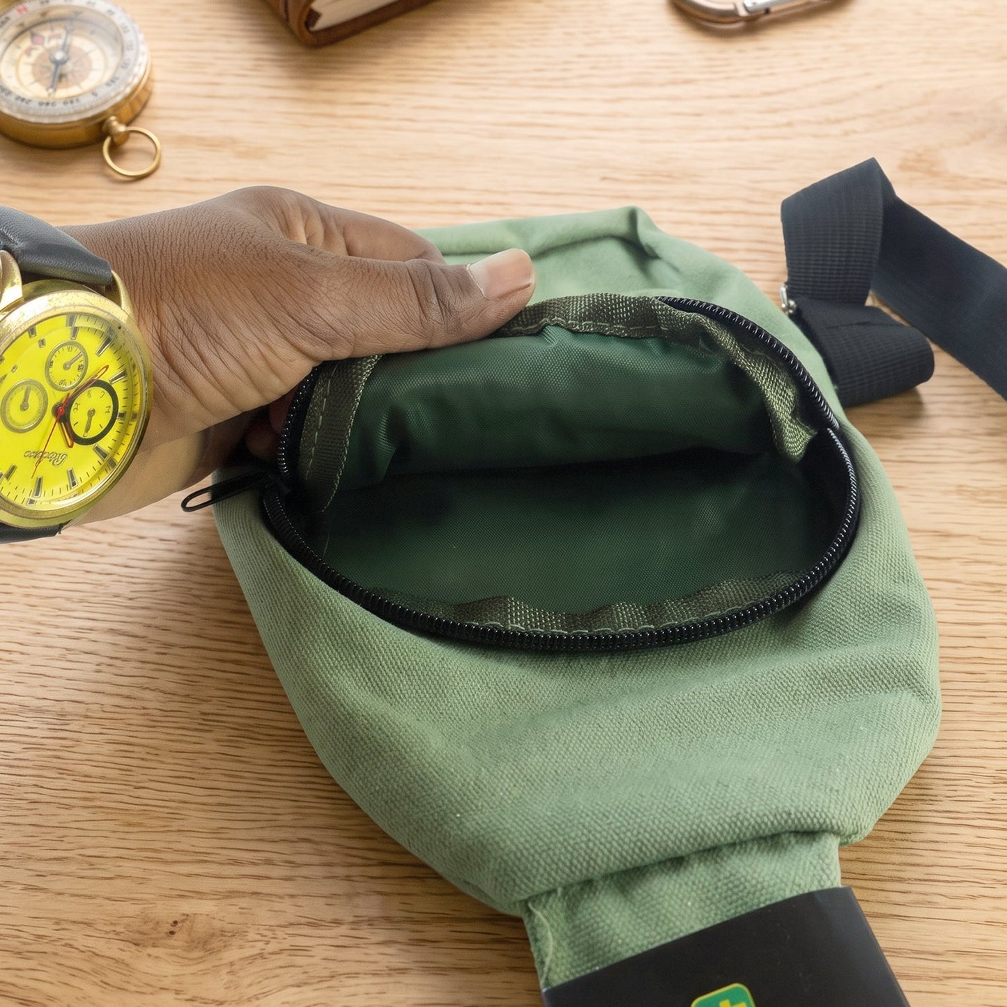 A hand opening the main zipper compartment of the green canvas sling bag, revealing a dark green interior lining.