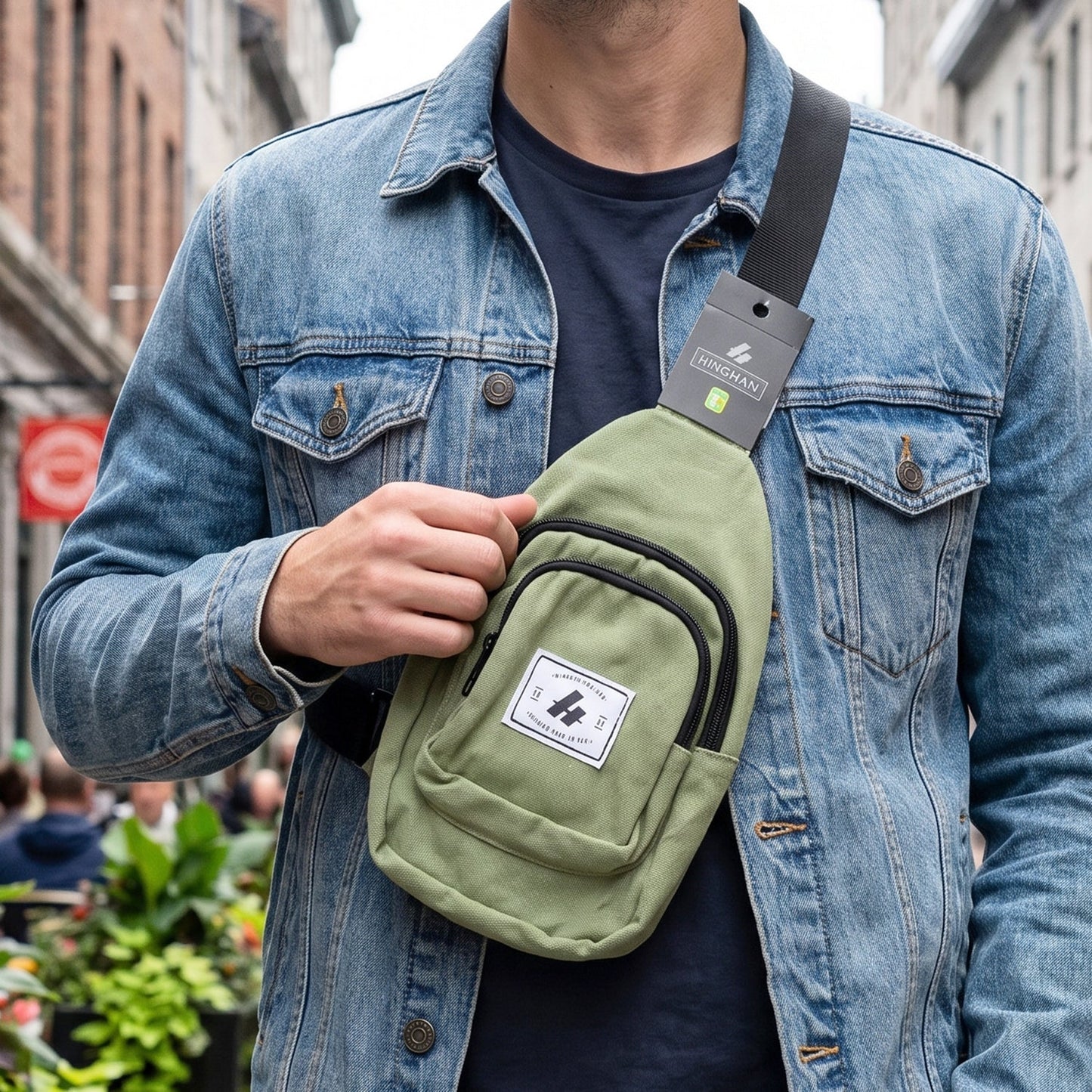 A man in a denim jacket wears a green canvas sling bag across his chest while walking outdoors.