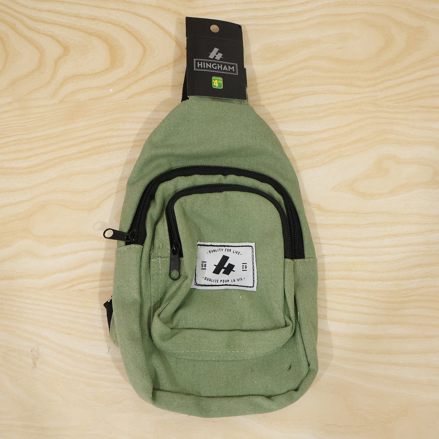 A green canvas sling bag with a black strap and zippers, featuring a white label, displayed flat on a light wood surface.