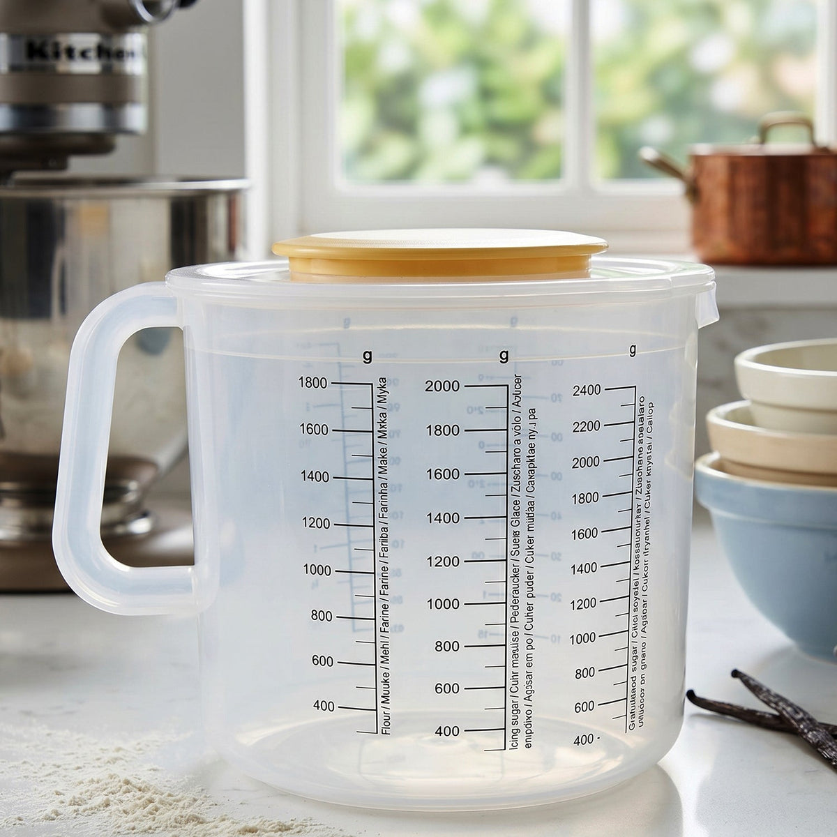 A clear plastic measuring jug with a yellow lid, positioned on a kitchen counter next to a stand mixer.