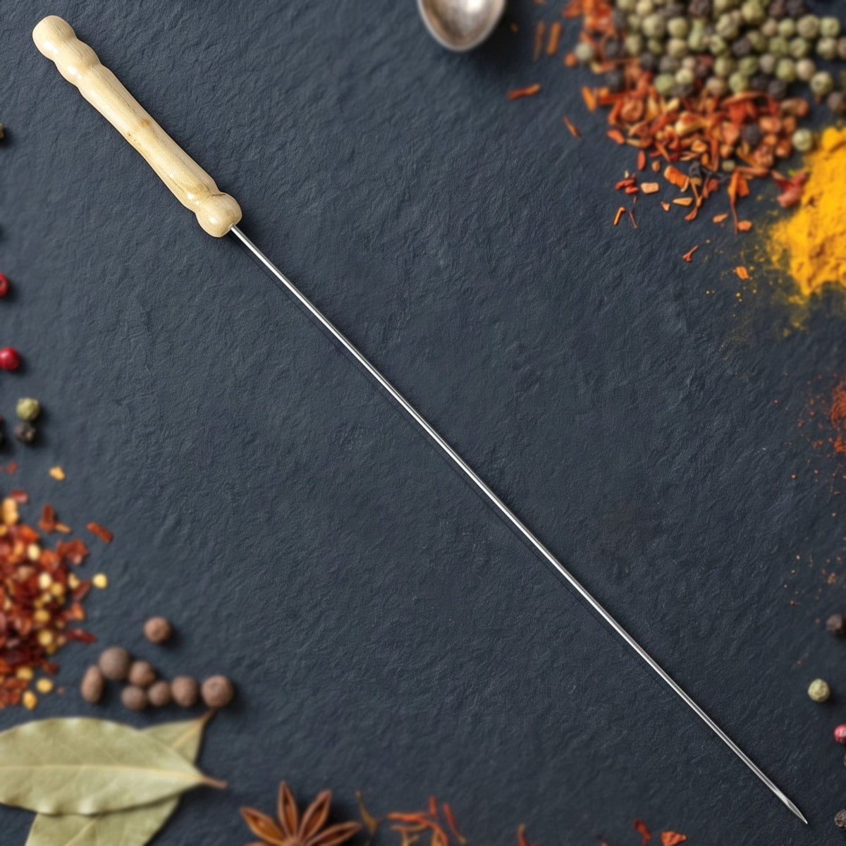 A single stainless steel skewer with a light wooden handle laid diagonally on a dark slate surface with scattered spices.
