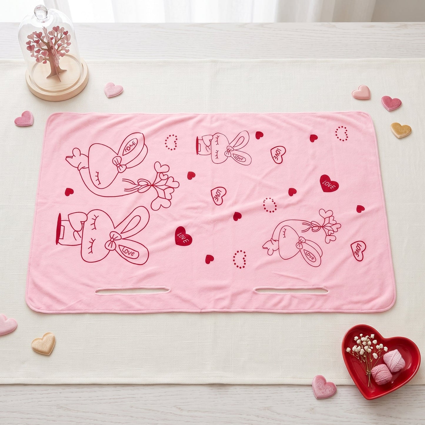 A pink towel with red bunny and heart prints lies on a white tablecloth with heart decorations.