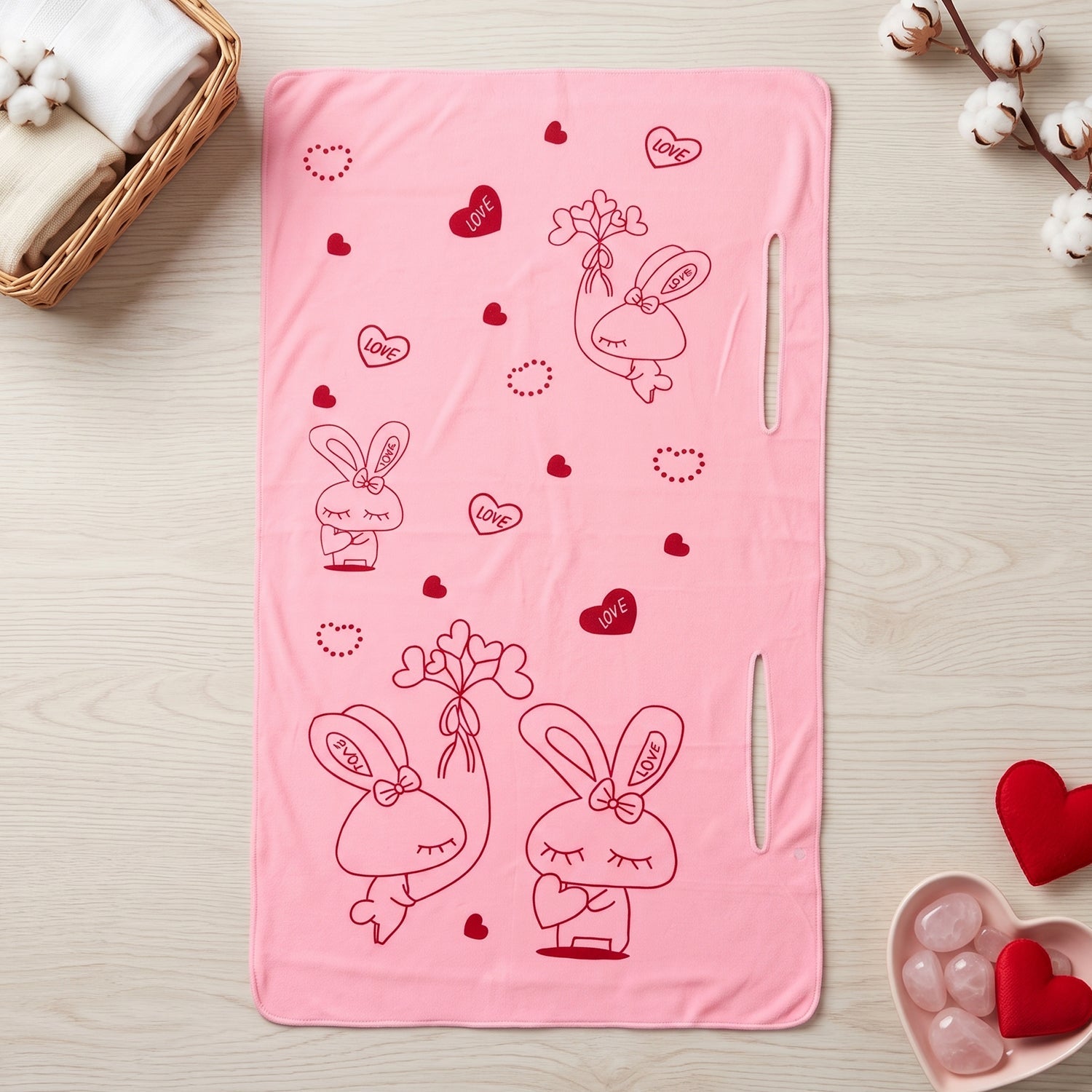 A pink towel with red bunny and heart prints lies flat on a wooden surface with cotton branches.
