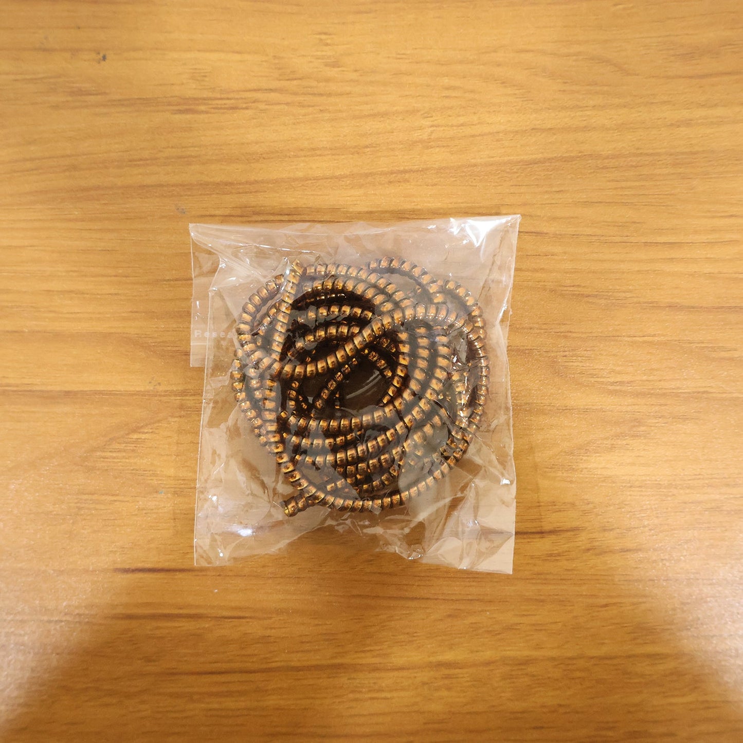 A coiled brown spiral cable protector sleeve is sealed inside a clear plastic bag on a wooden surface.