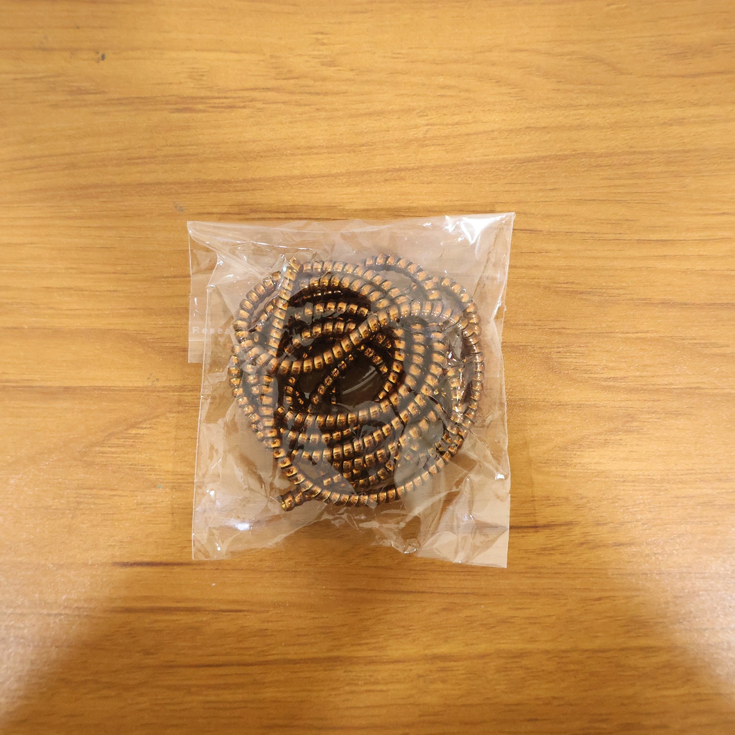A coiled brown spiral cable protector sleeve is sealed inside a clear plastic bag on a wooden surface.