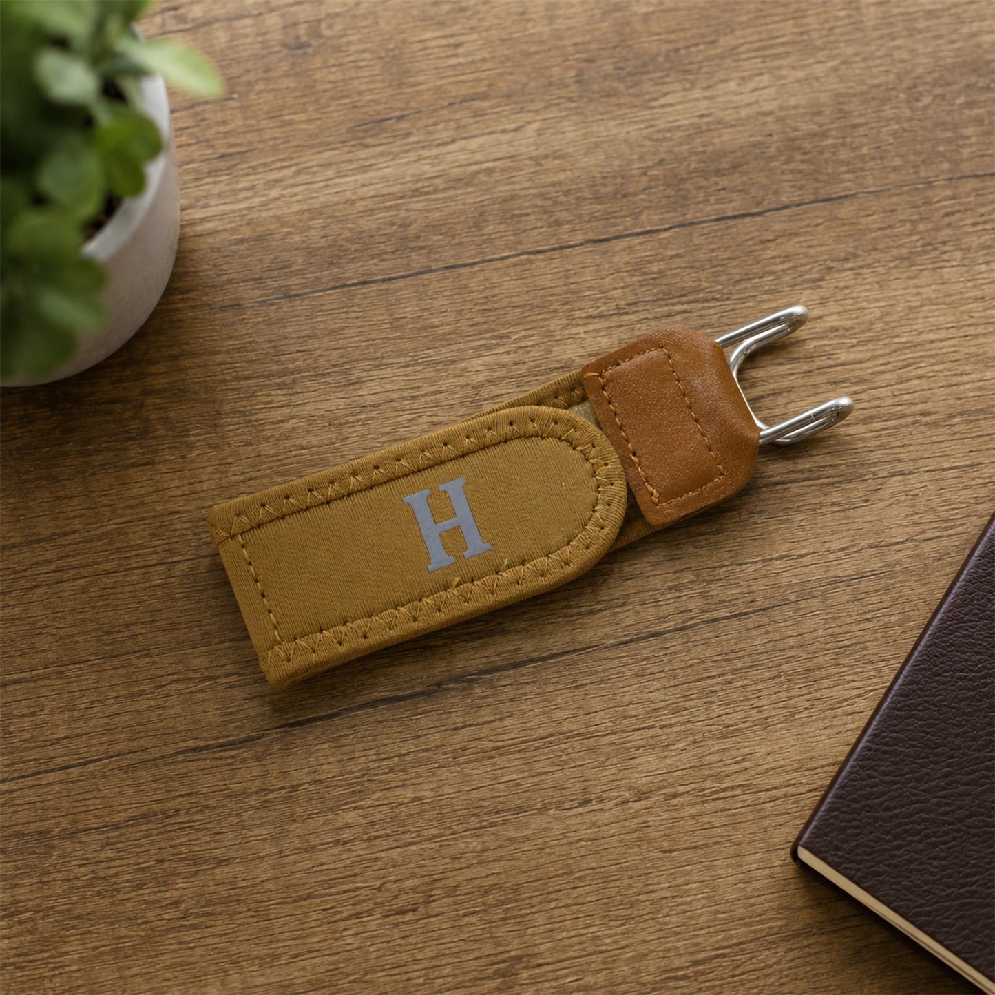 The camel elastic belt with a gray 'H' and silver hooks rests on a wooden surface next to a small green plant and a notebook.