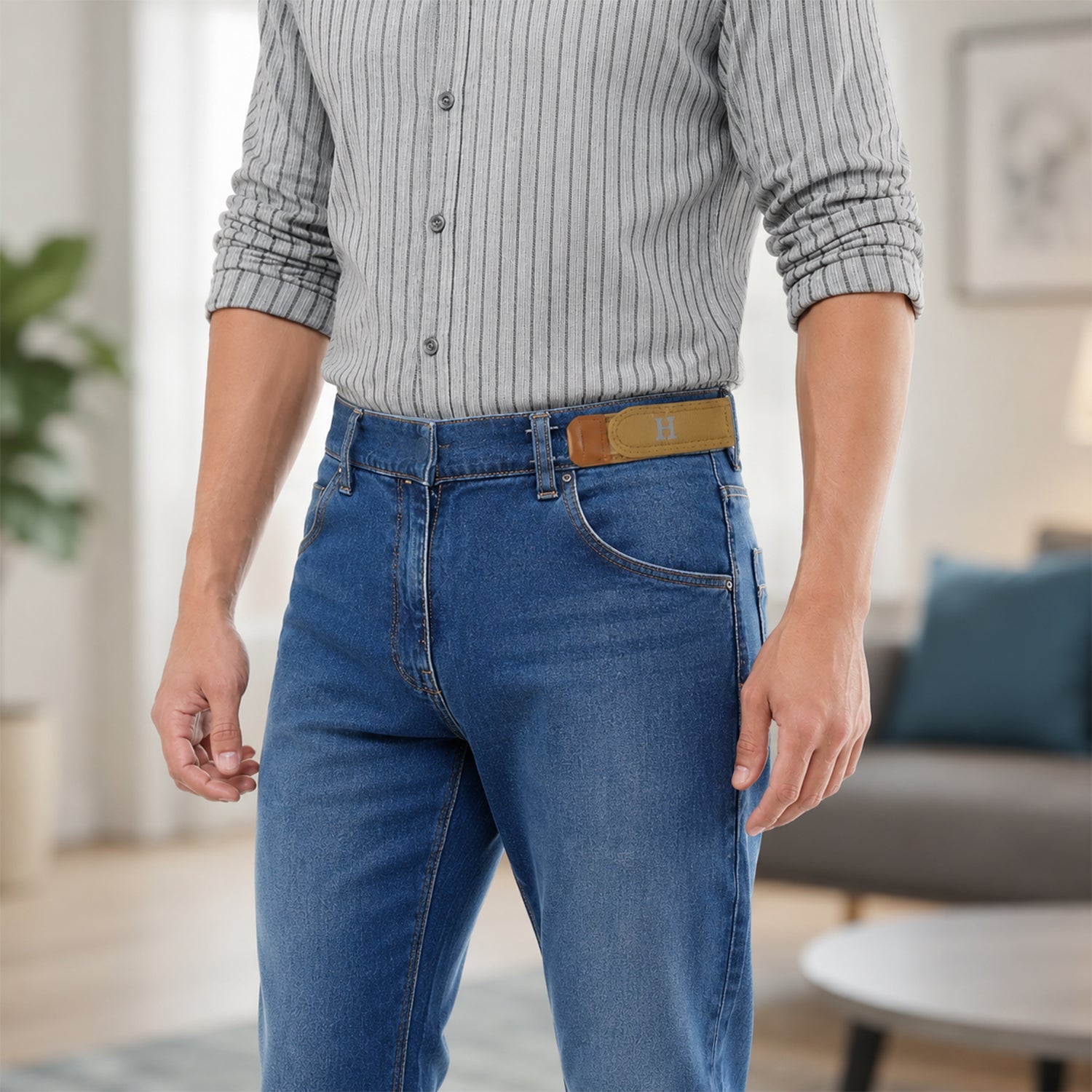 A man in a striped shirt and blue jeans wears a tan buckle-less belt with a silver 'H' on the front.