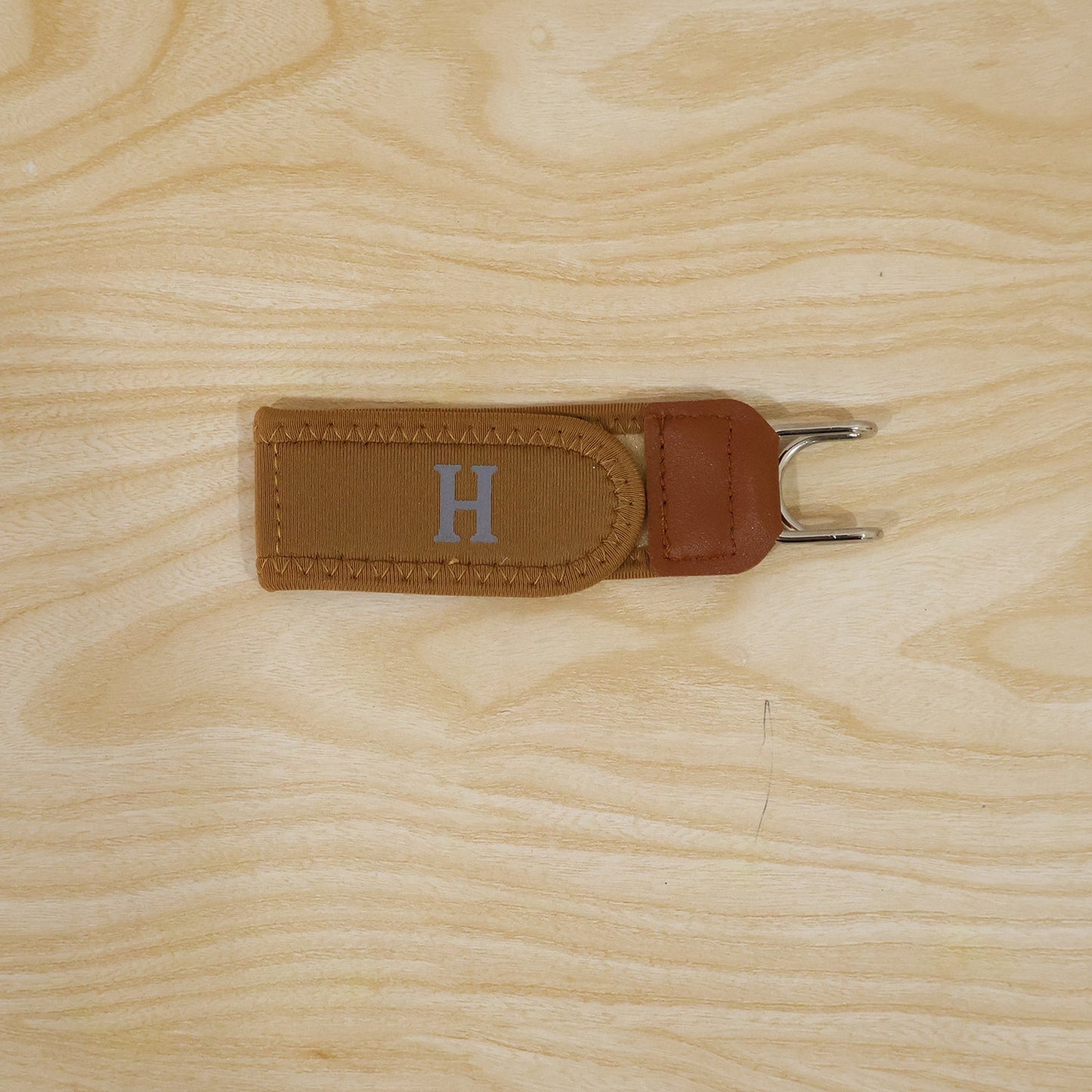 A tan buckle-less belt with a silver 'H' is centered on a light wooden surface.
