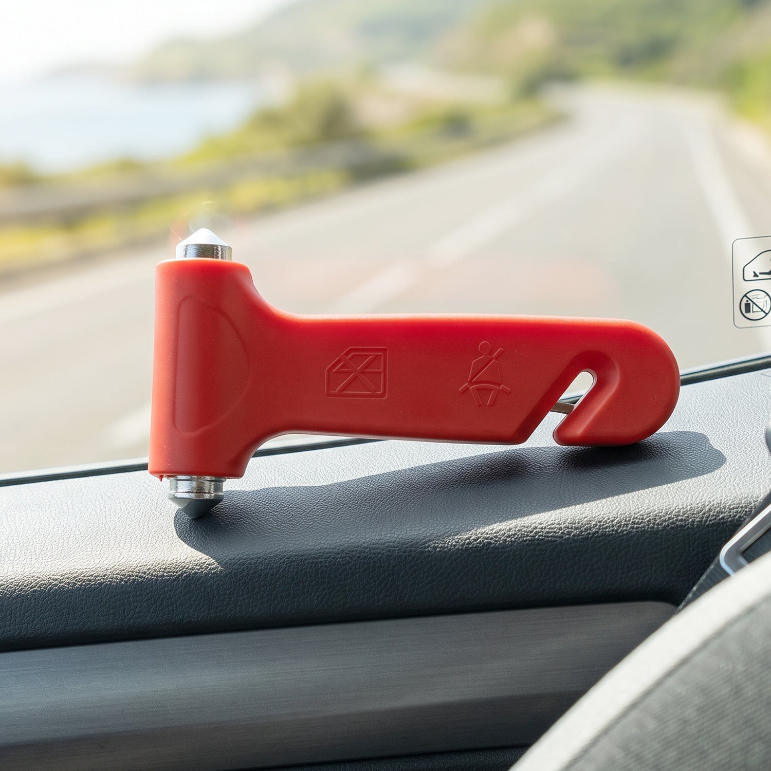 A red car safety hammer with safety icons rests on a car dashboard, with a road and ocean in the background.