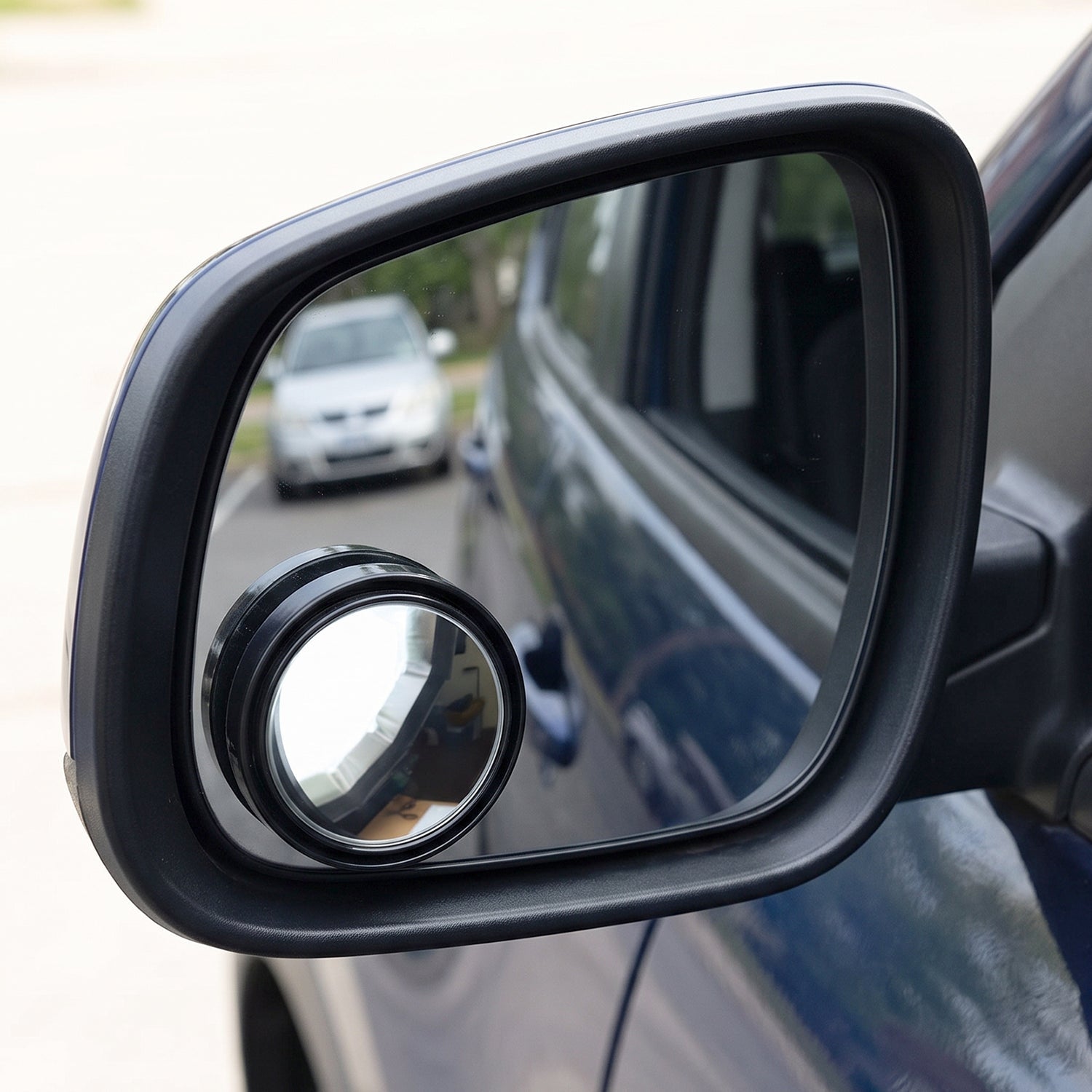 A black car side mirror with a round blind spot mirror attached, showing a white car in the reflection.