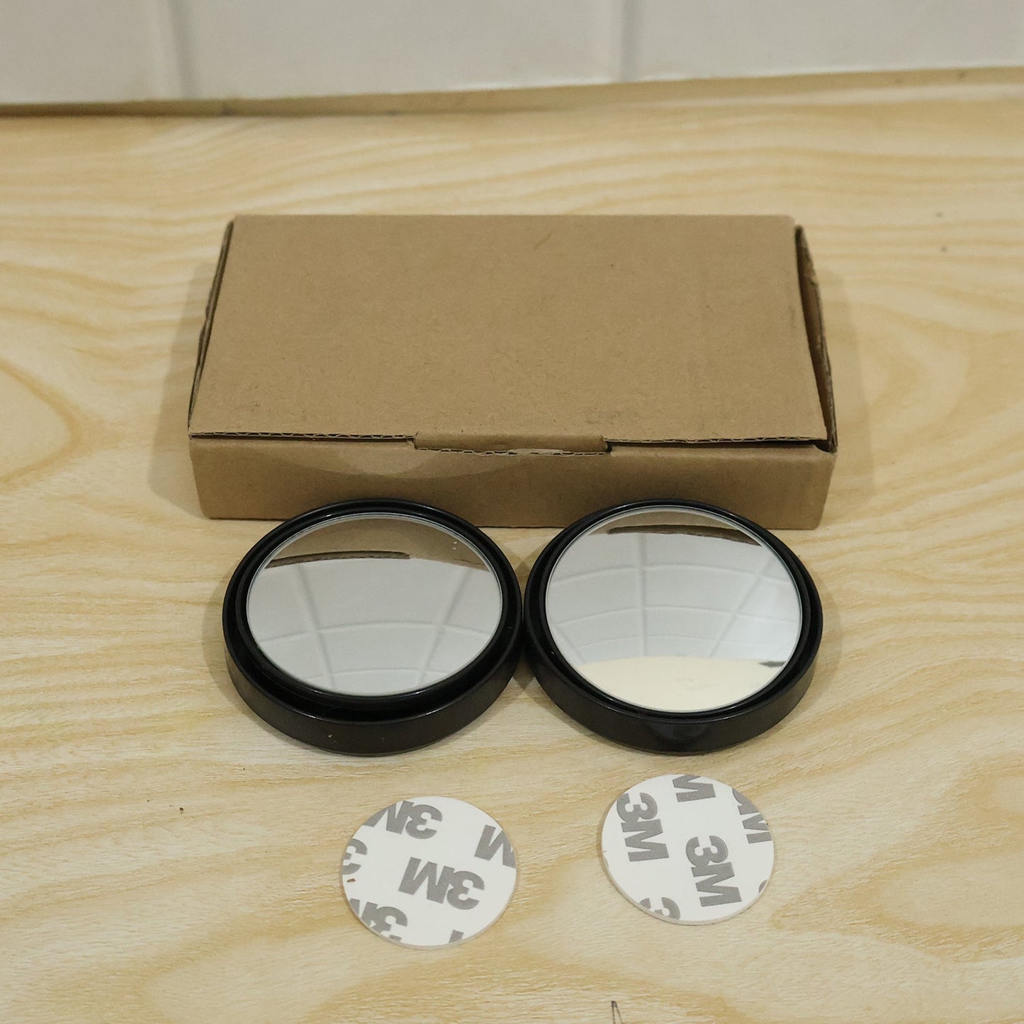 Two round blind spot mirrors with black frames, two 3M adhesive pads, and a brown cardboard box on a wooden surface.