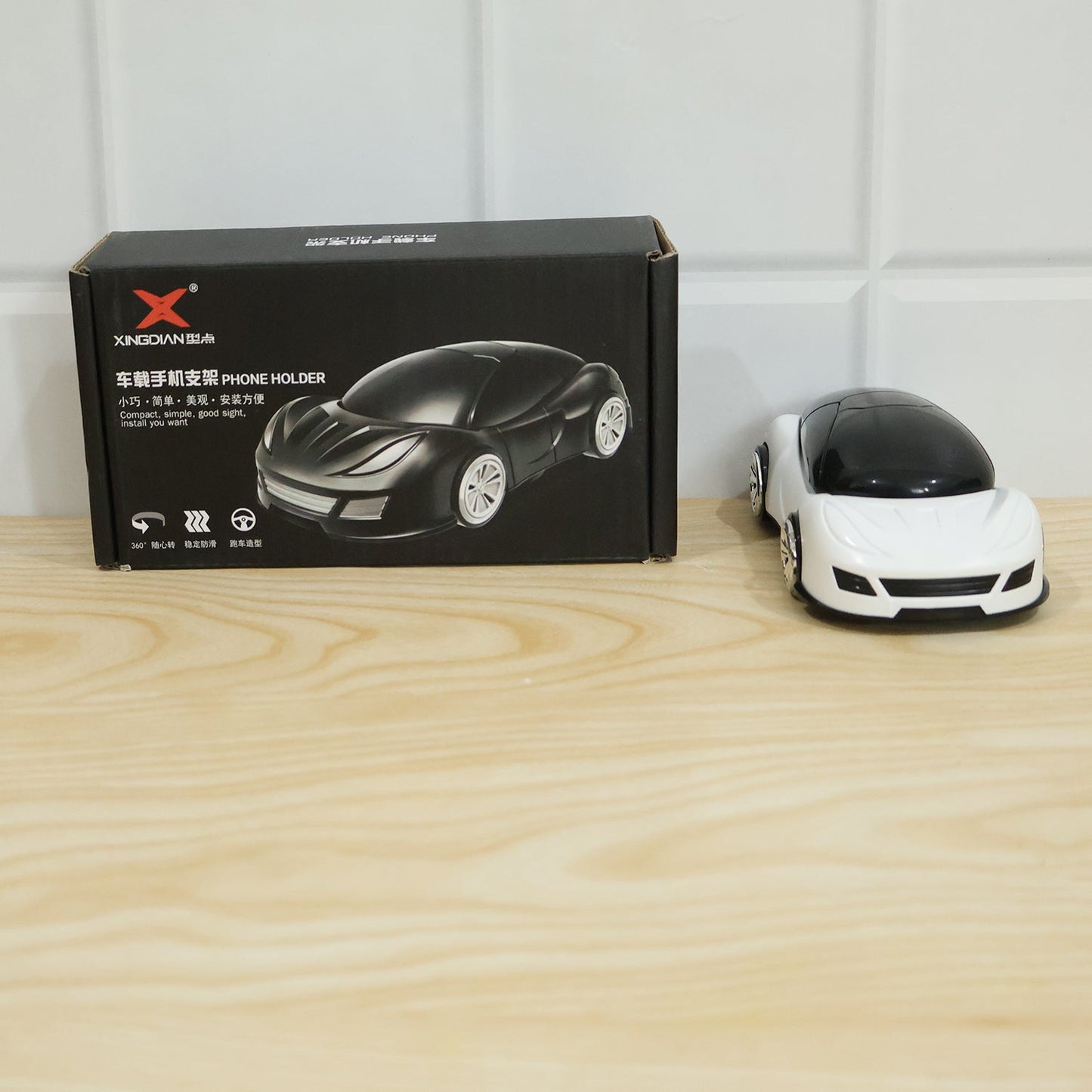 A black box labeled 'XINGDIAN PHONE HOLDER' sits next to a white car-shaped phone holder on a wooden surface.