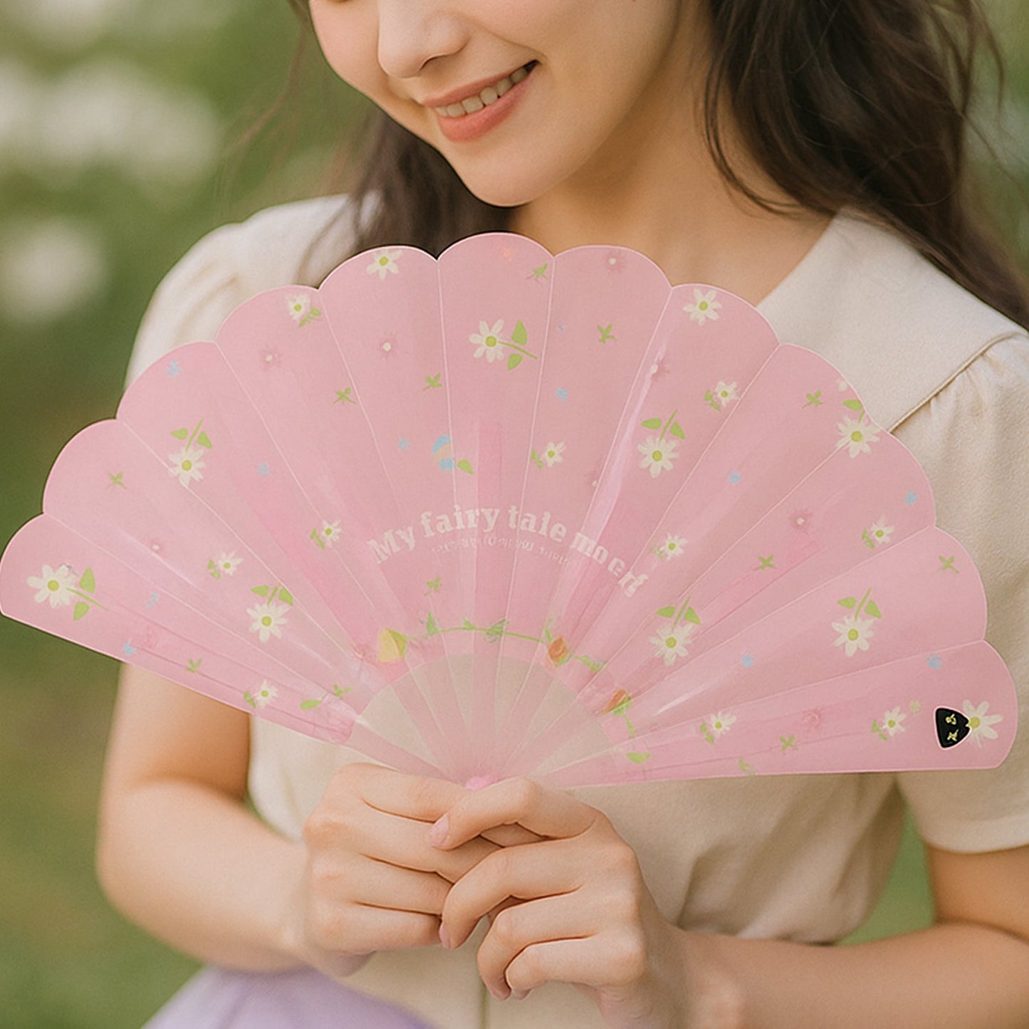 A woman holds an open pink fan with white floral patterns and the text