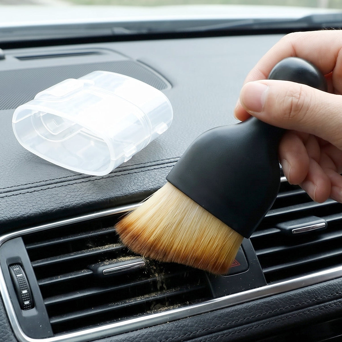 A black brush with light brown bristles cleaning dust from a car's dashboard vent, with a clear plastic cover beside it.
