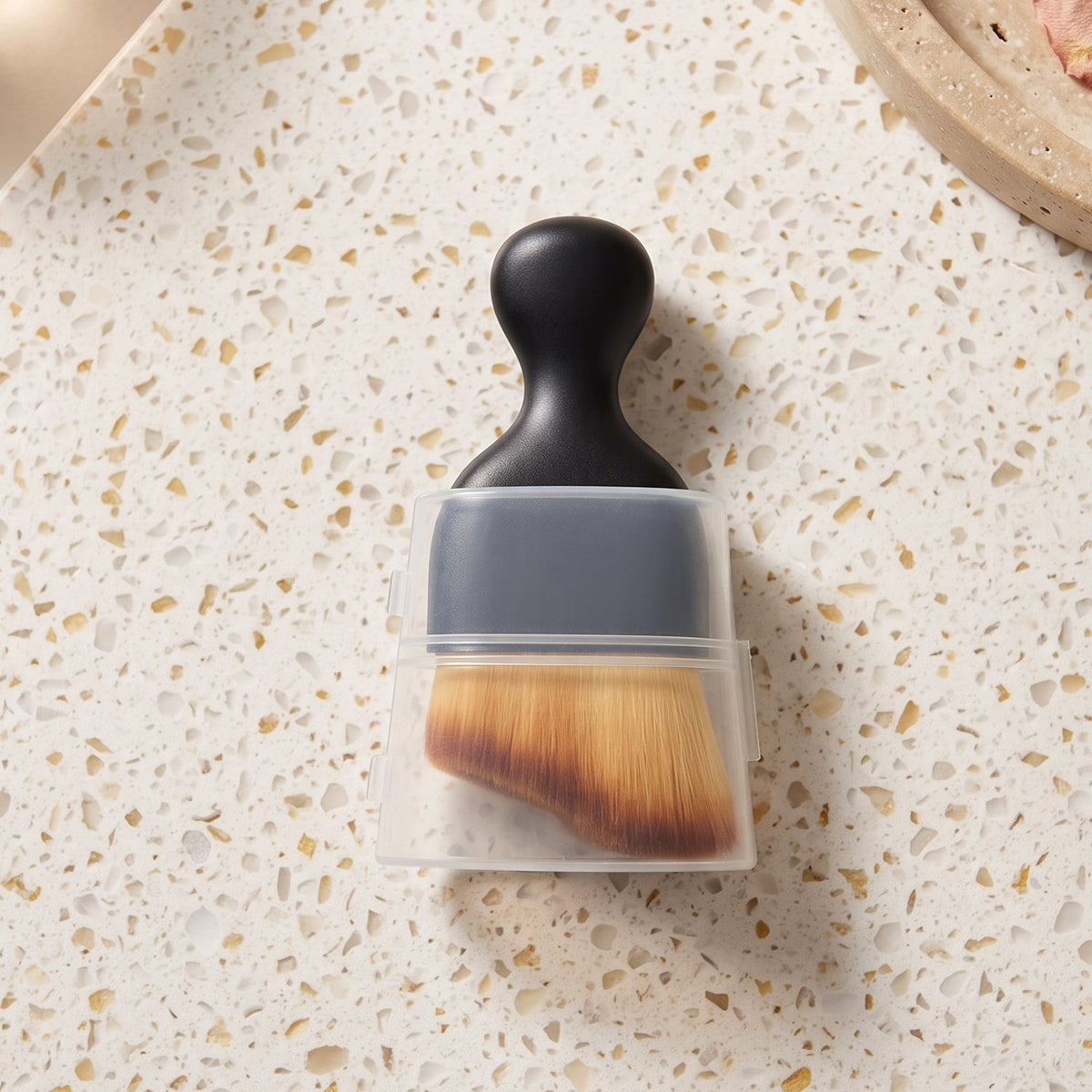 A black-handled brush with light brown bristles, partially covered by a clear plastic case, resting on a speckled white