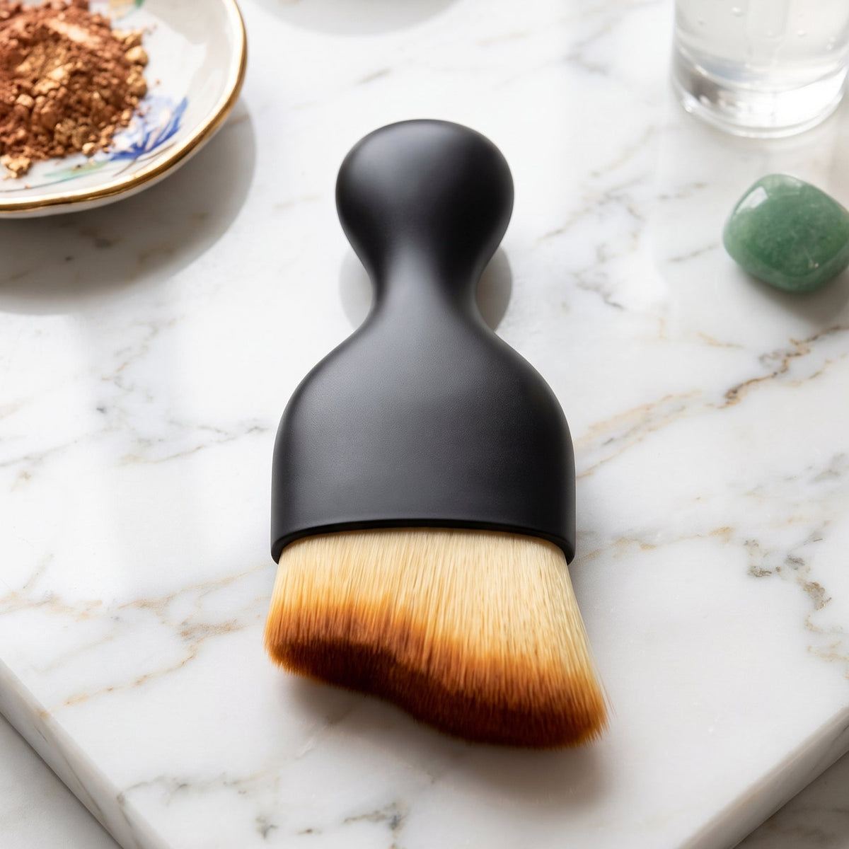 A black-handled brush with light brown, angled bristles resting on a white marble surface, with a small dish of powder in