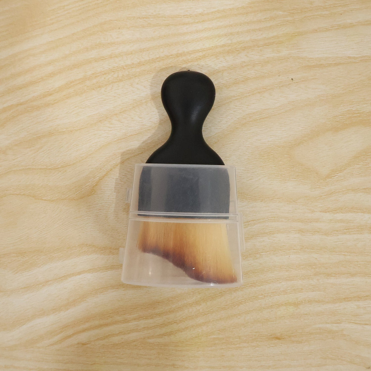 A black-handled brush with light brown bristles, partially covered by a clear plastic case, on a light wooden surface.