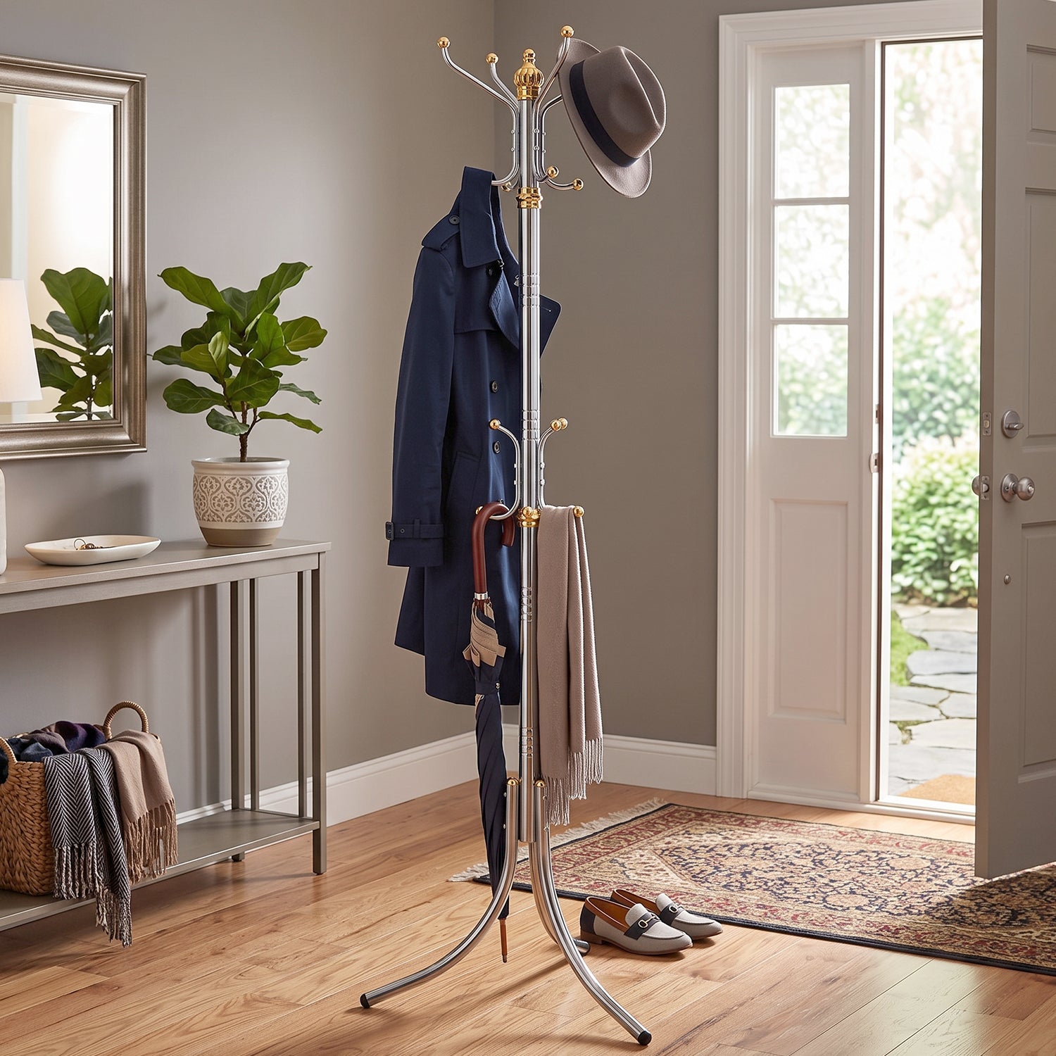 A silver and gold metal stand holds a blue coat, a brown hat, an umbrella, and a scarf in an entryway with a plant, mirror,