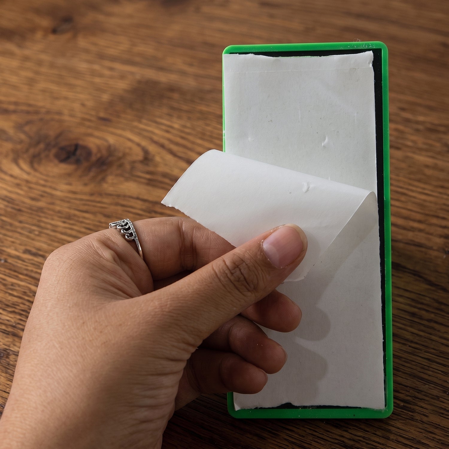 A hand with a silver ring peels the protective backing off the adhesive base of a green knife sharpener.