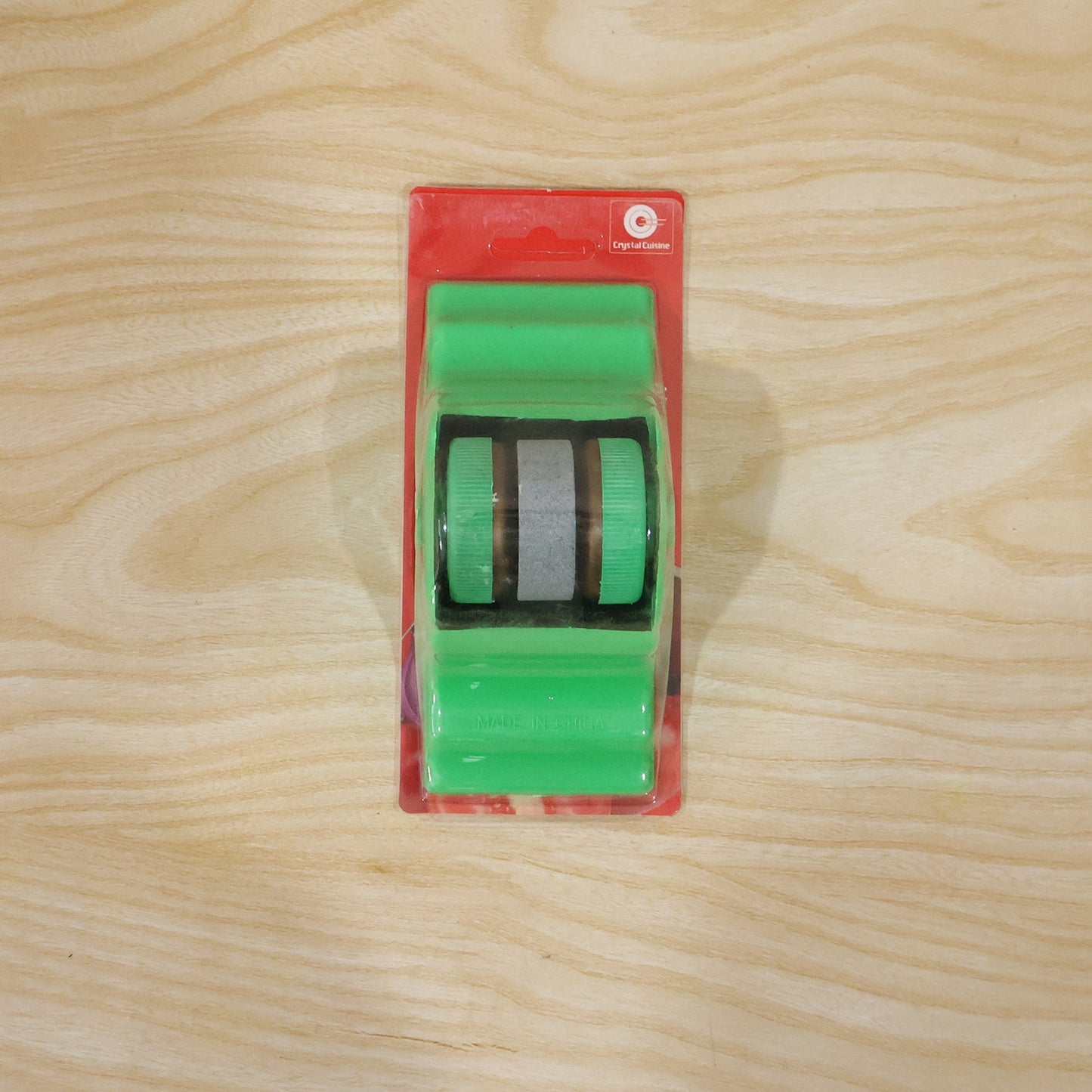 A green compact knife sharpener is sealed in clear plastic packaging with a red cardboard backing on a wooden surface.