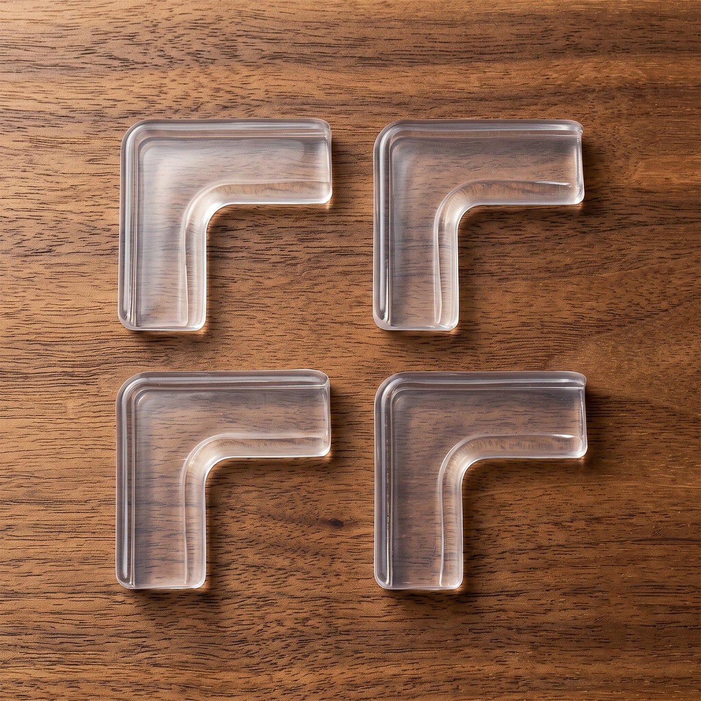 Four clear silicone corner protectors are arranged in a grid on a dark brown wooden surface, showcasing their L-shape.