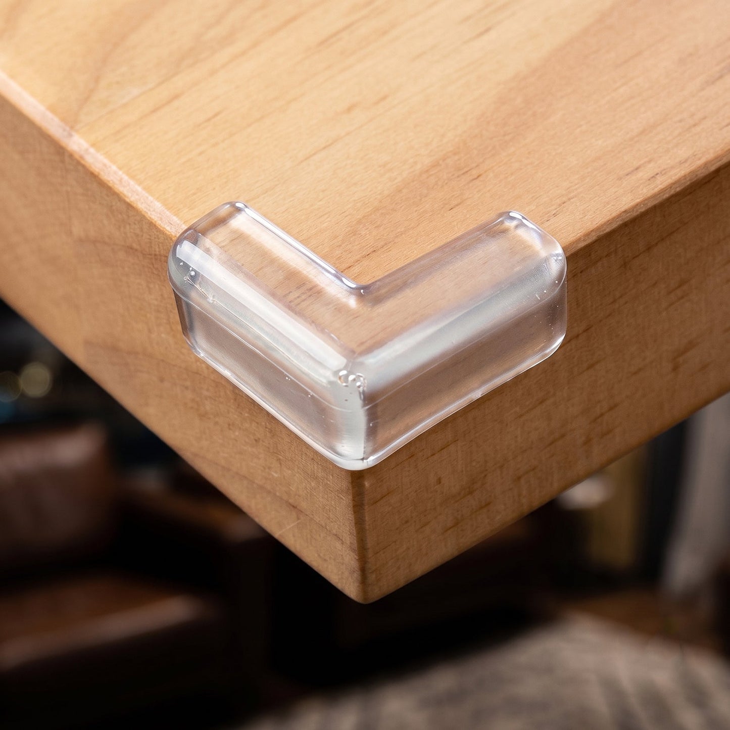 A clear silicone corner protector is securely placed on the corner of a light brown wooden table.