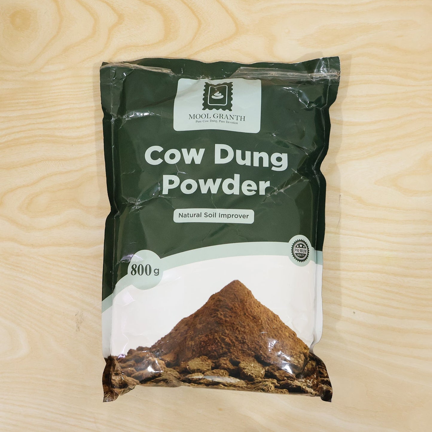 A single dark green bag of "Cow Dung Powder" with a brown mound illustration is centered on a light wooden background.