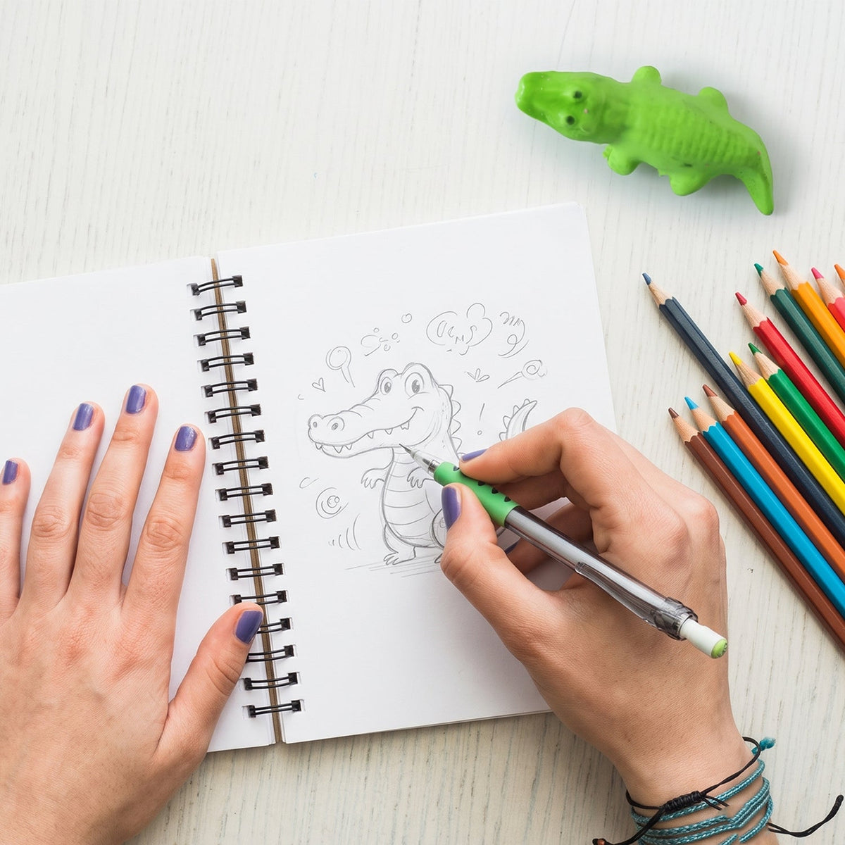 A person's hands draw a cartoon crocodile in a spiral-bound notebook, with a green crocodile eraser and colored pencils on