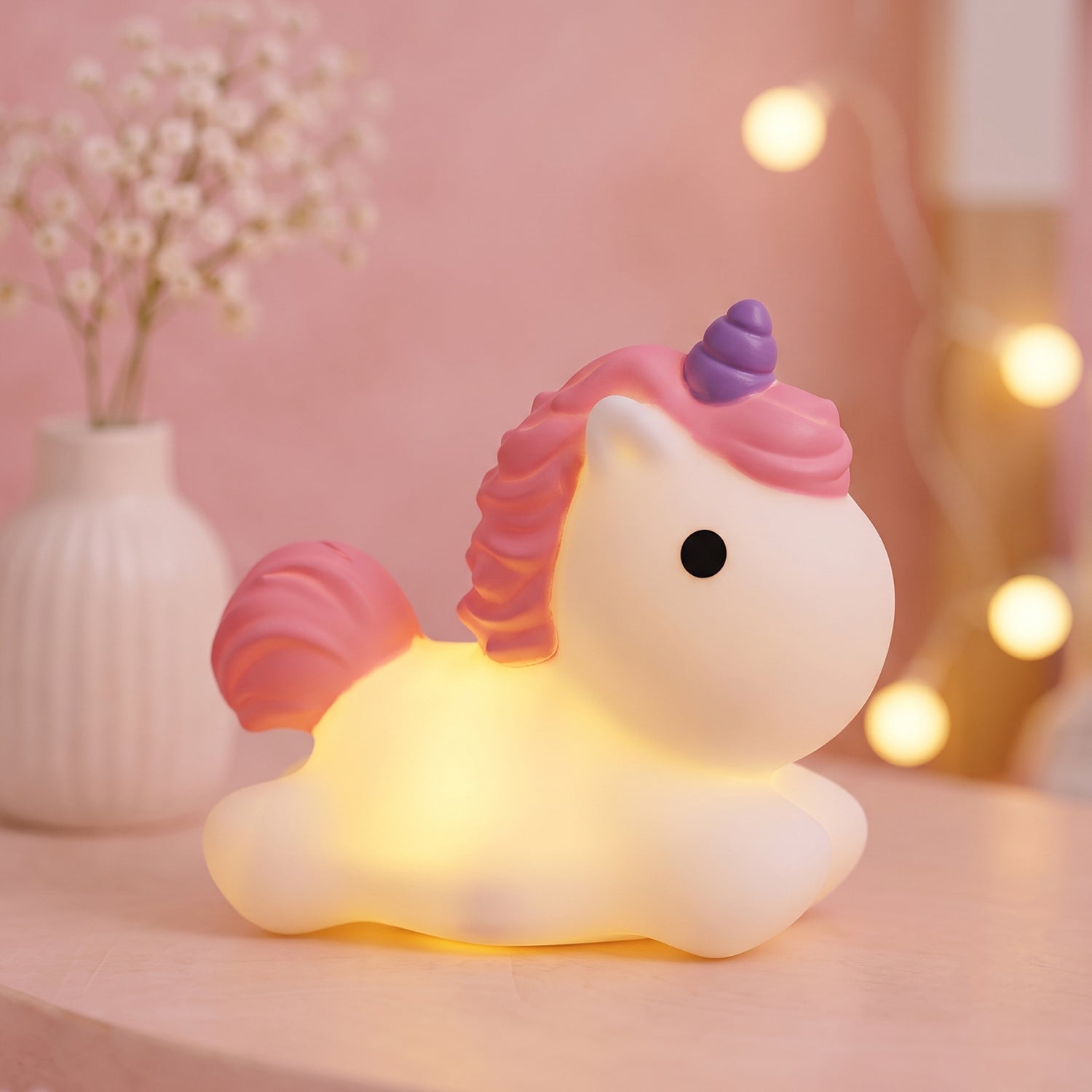 A glowing white unicorn night light with pink mane and tail, and a purple horn, sits on a pink surface next to a white vase