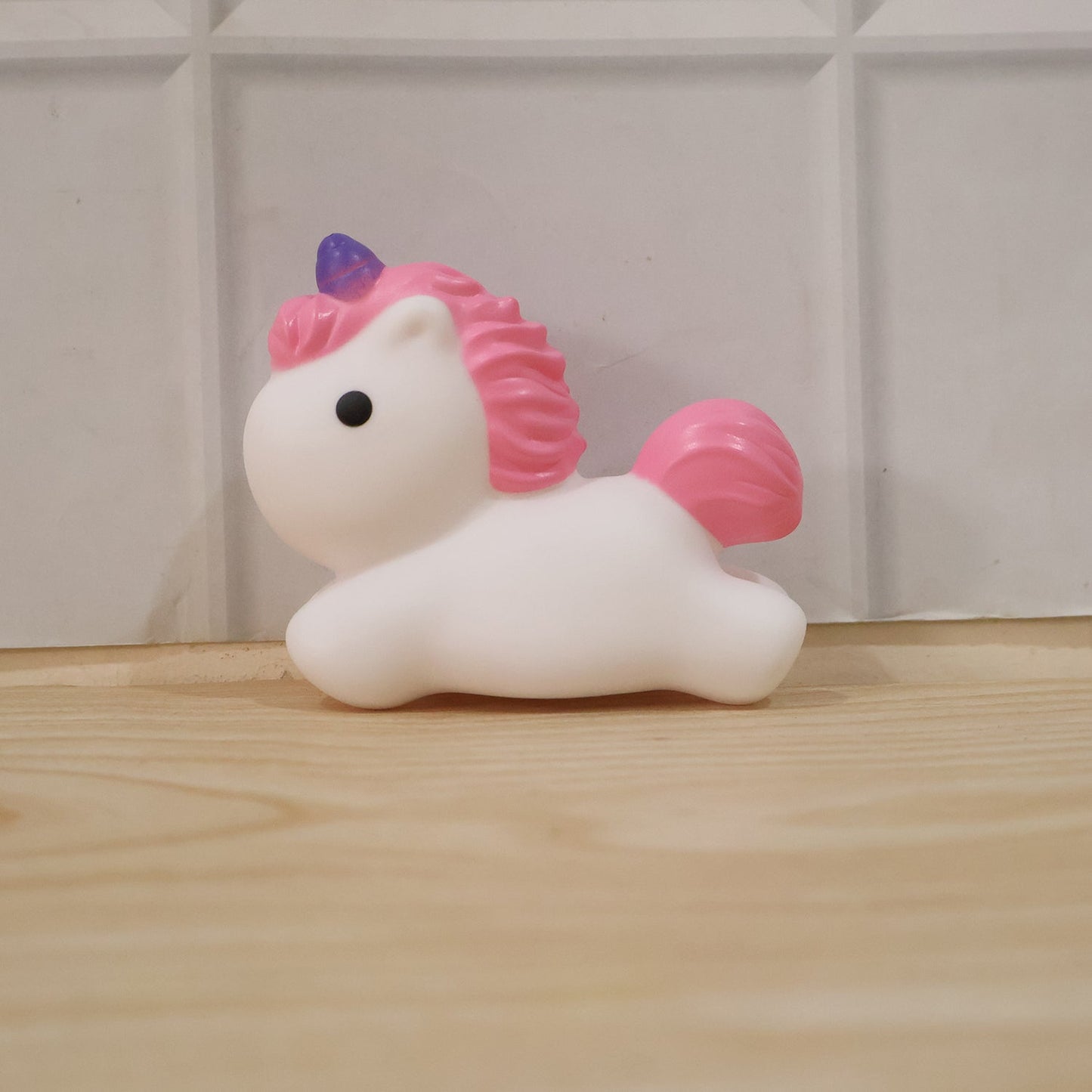 An unlit white unicorn with pink mane and tail, and a purple horn, sits on a light wooden floor against a white tiled wall.