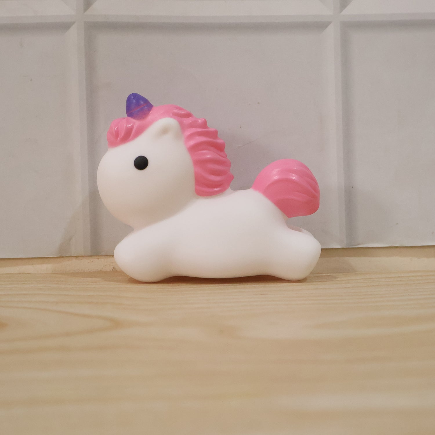 An unlit white unicorn with pink mane and tail, and a purple horn, sits on a light wooden floor against a white tiled wall.