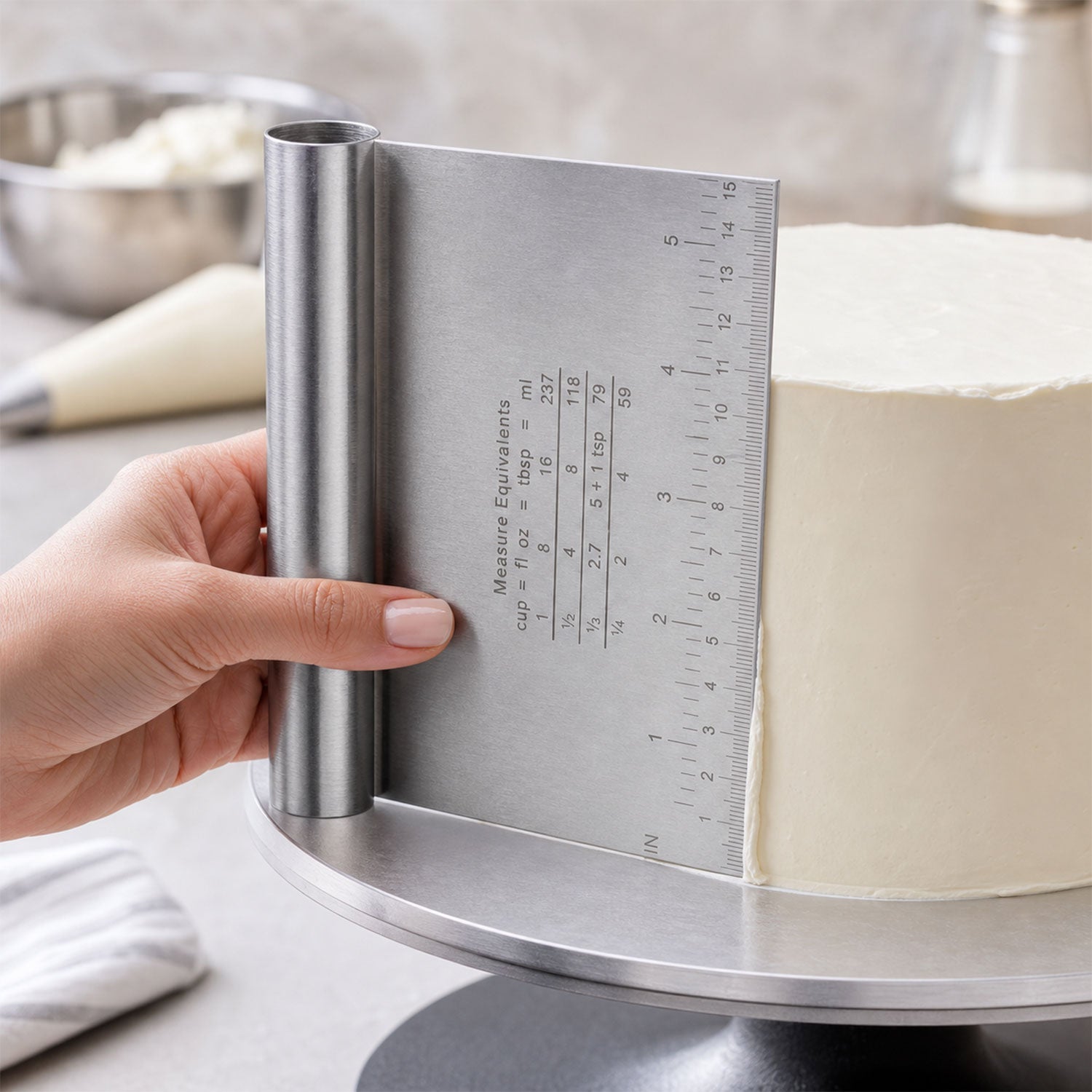 A hand uses a stainless steel dough scraper to smooth the side of a white frosted cake on a rotating cake stand.