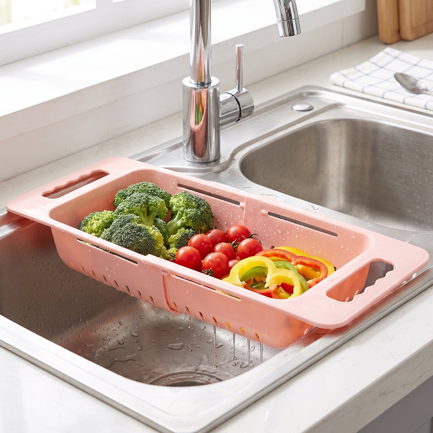 Pink plastic adjustable over-sink drain basket with broccoli, tomatoes, and bell peppers in a kitchen sink, with a checkered