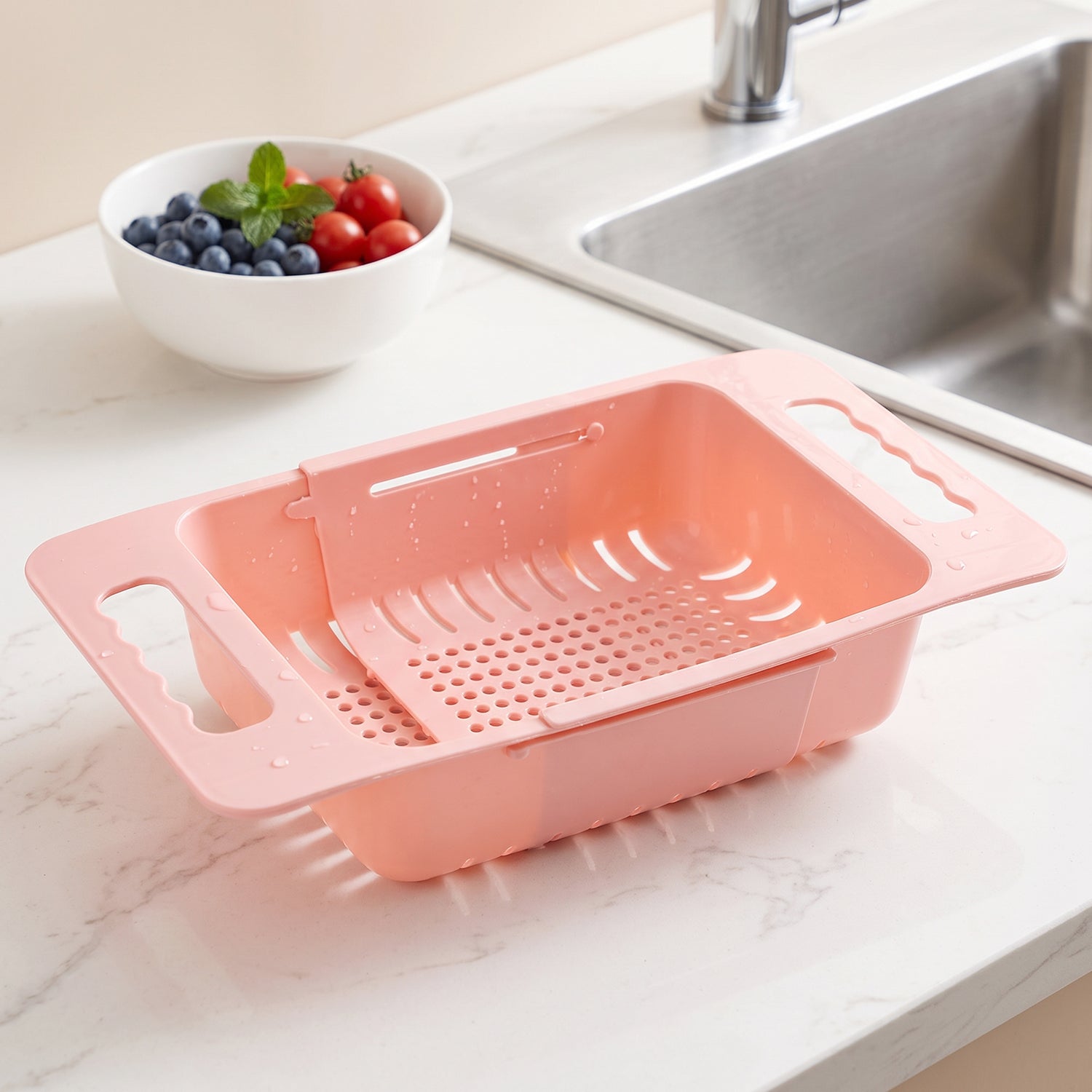 Pink plastic adjustable over-sink drain basket on a white countertop with a bowl of berries and a sink in the background.