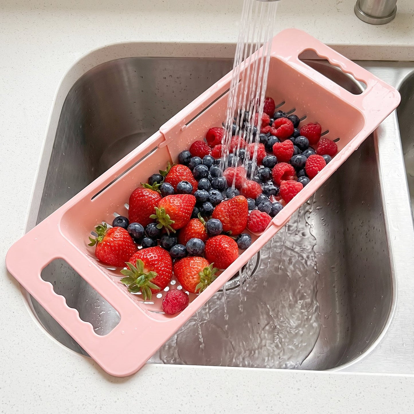 Pink plastic adjustable over-sink drain basket filled with strawberries, blueberries
