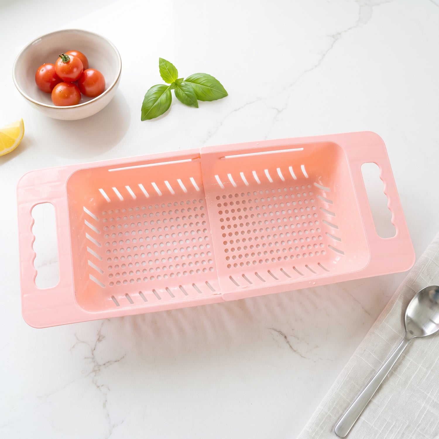 Pink plastic adjustable over-sink drain basket on a white marble countertop with a bowl of tomatoes and basil.