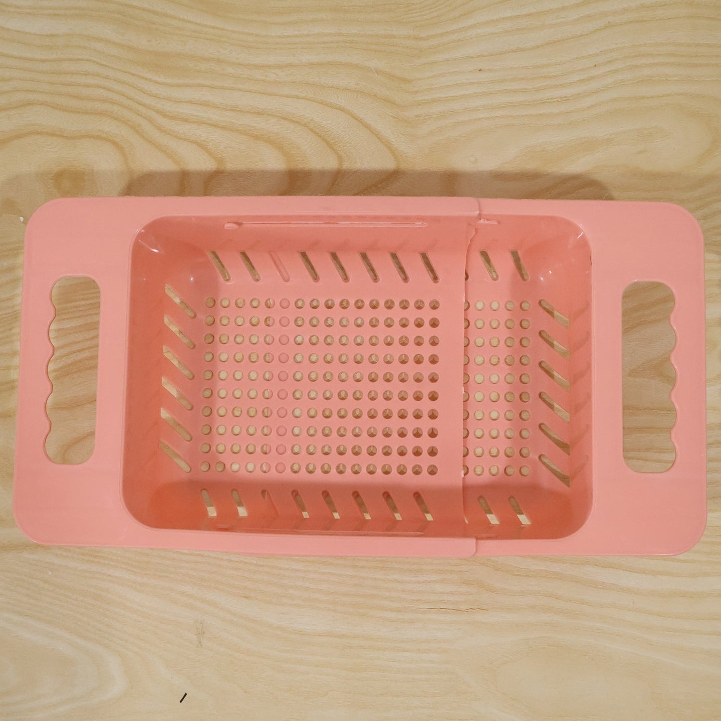 Pink plastic adjustable over-sink drain basket with perforated bottom and handles, extended on a light wooden surface.