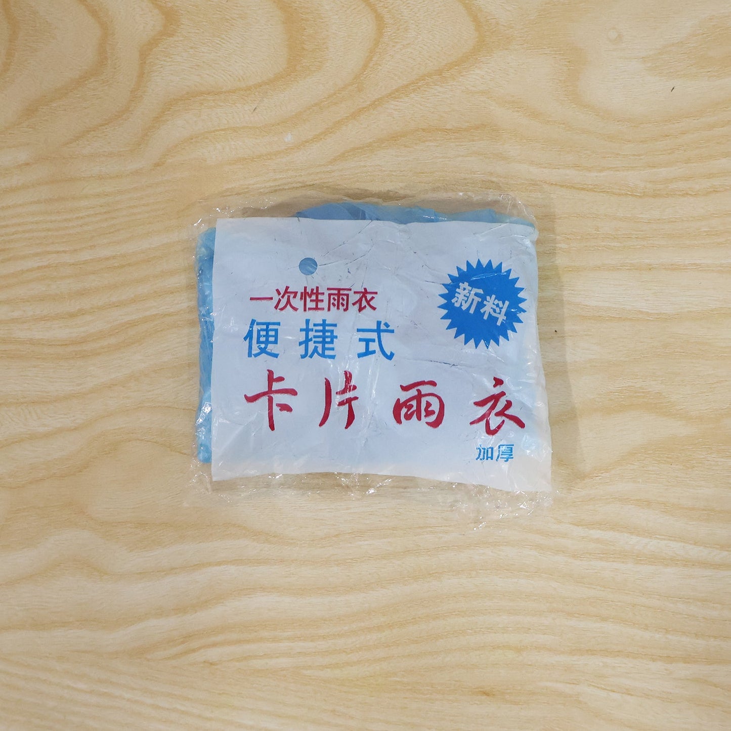 A small, clear plastic package containing a folded blue rain poncho is centered on a light wooden surface.